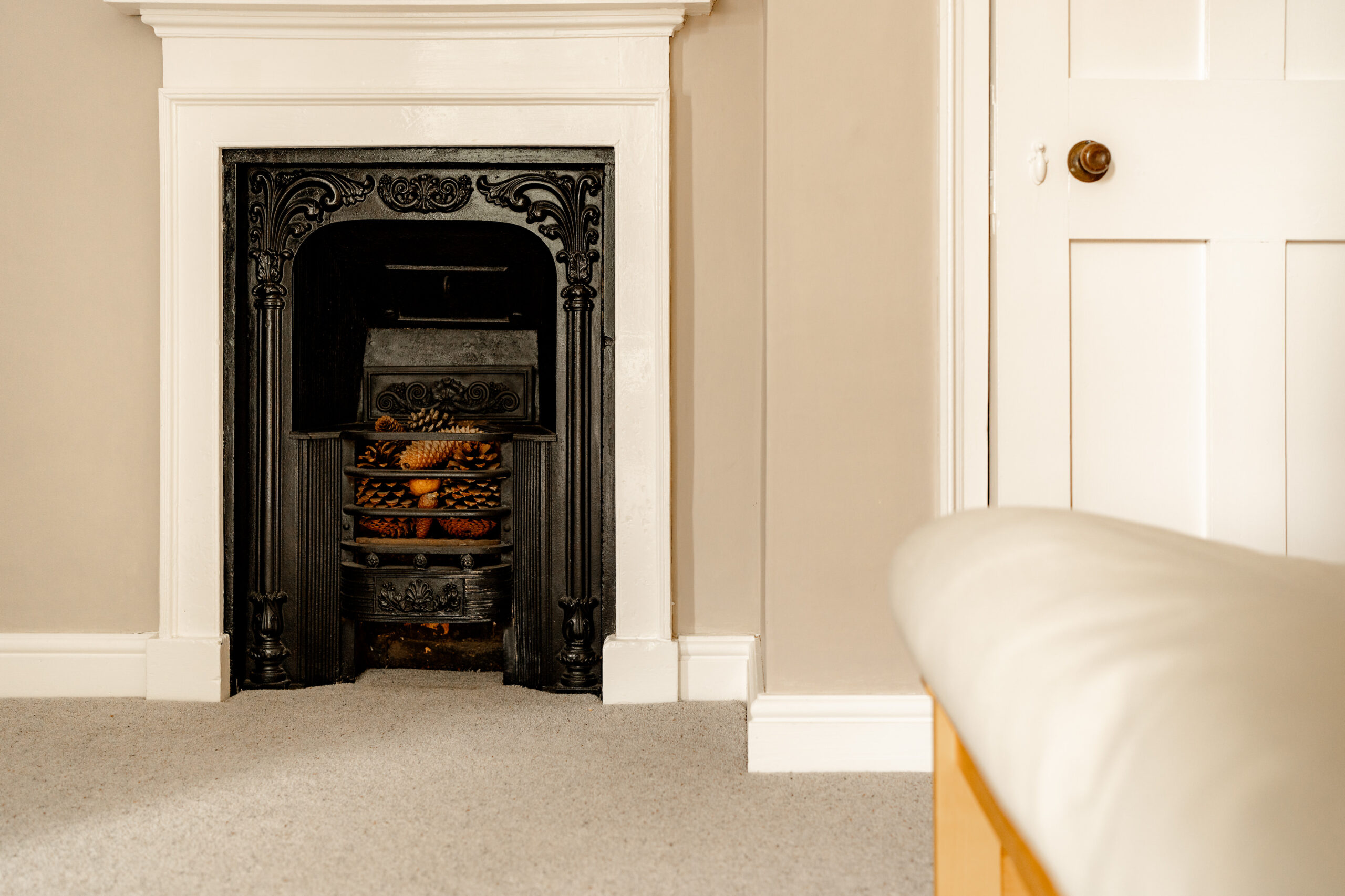 A black ornate fireplace with pinecones inside sits against a beige wall, next to a closed white door and a bed with a white comforter on a carpeted floor.