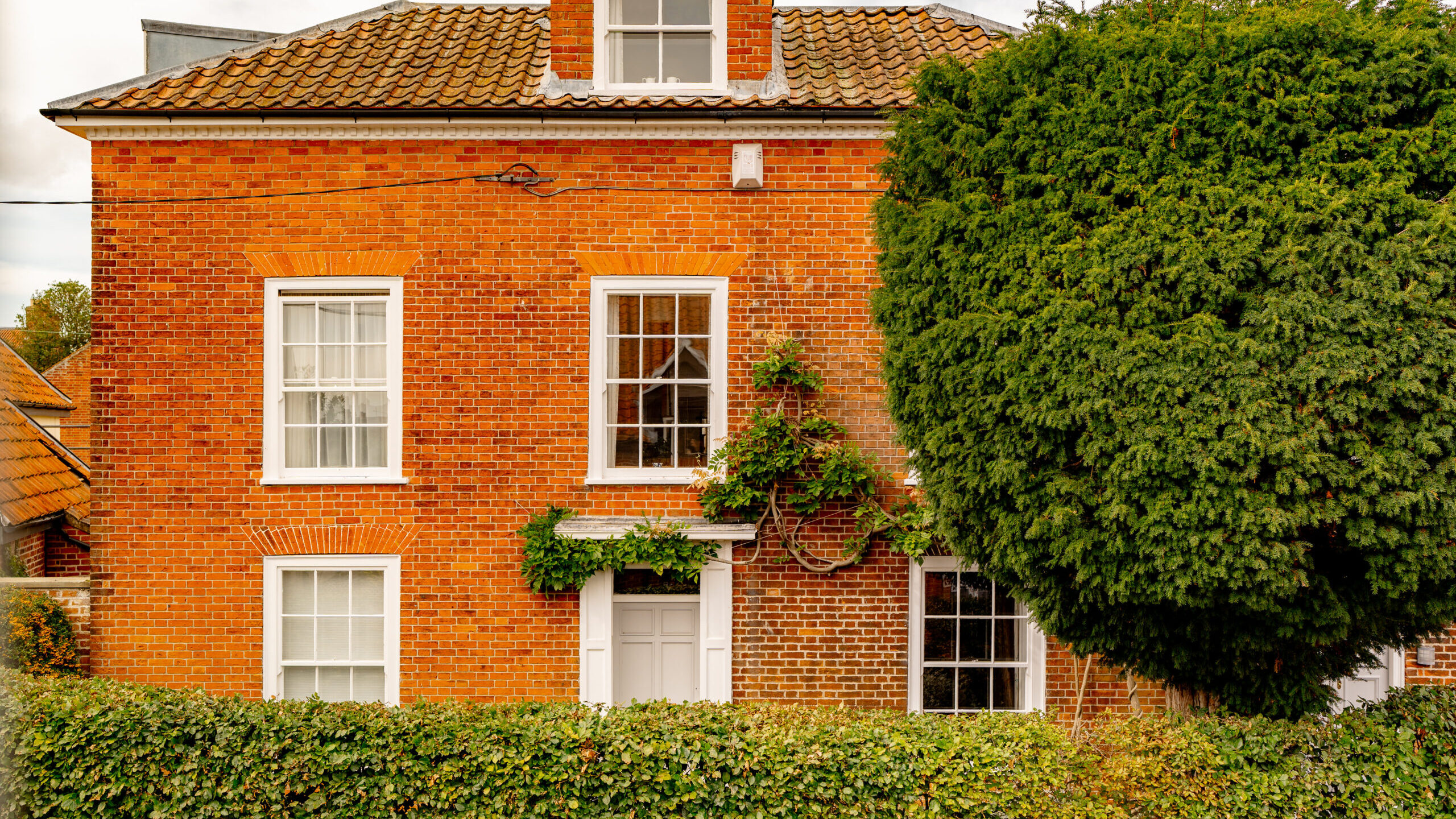 A red-brick house with white-framed windows, a tiled roof, and ivy growing on the wall. A tall, dense hedge and a large tree partially obscure the lower part of the building.