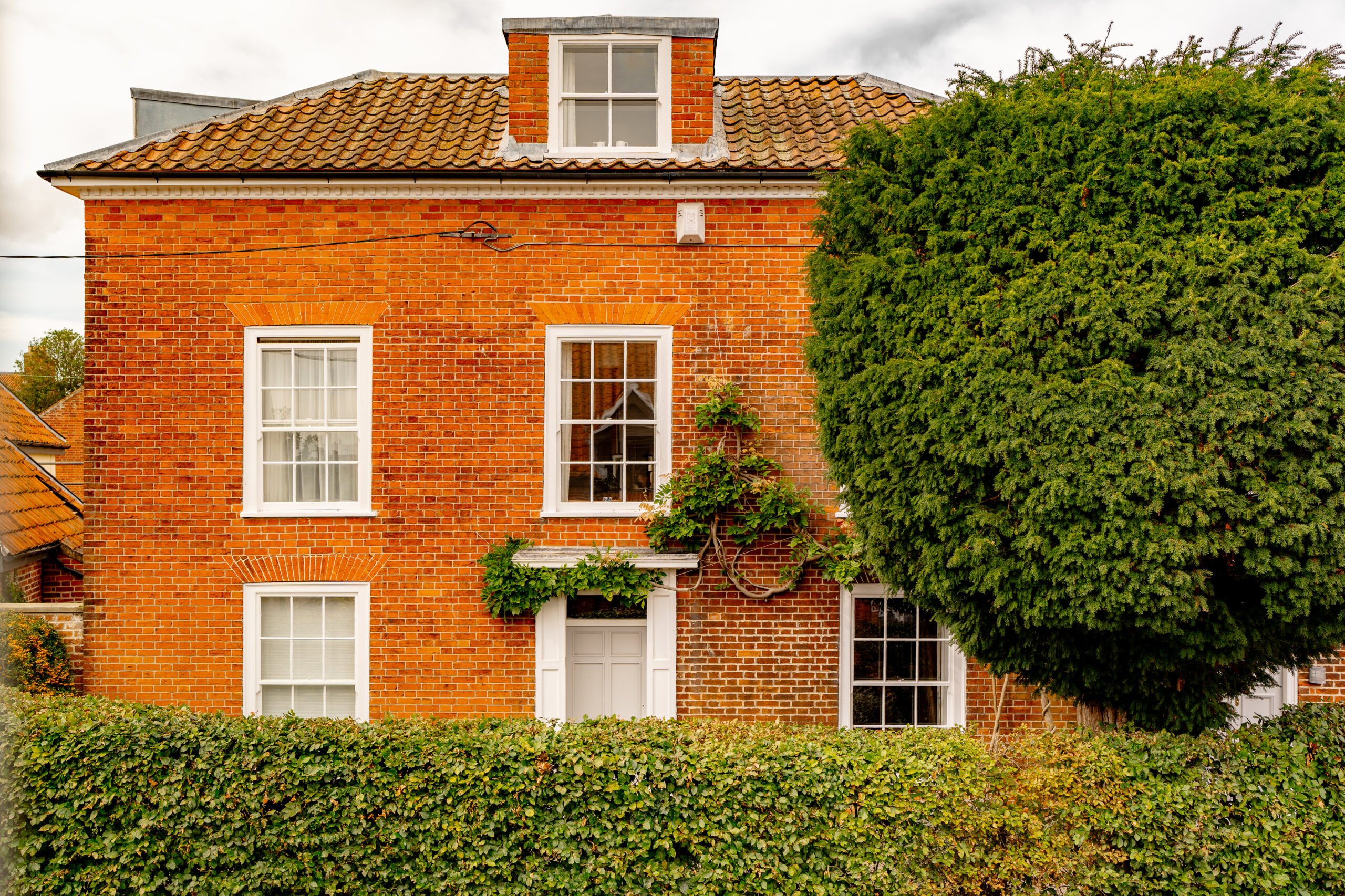 A red-brick house with white-framed windows, a tiled roof, and ivy growing on the wall. A tall, dense hedge and a large tree partially obscure the lower part of the building.