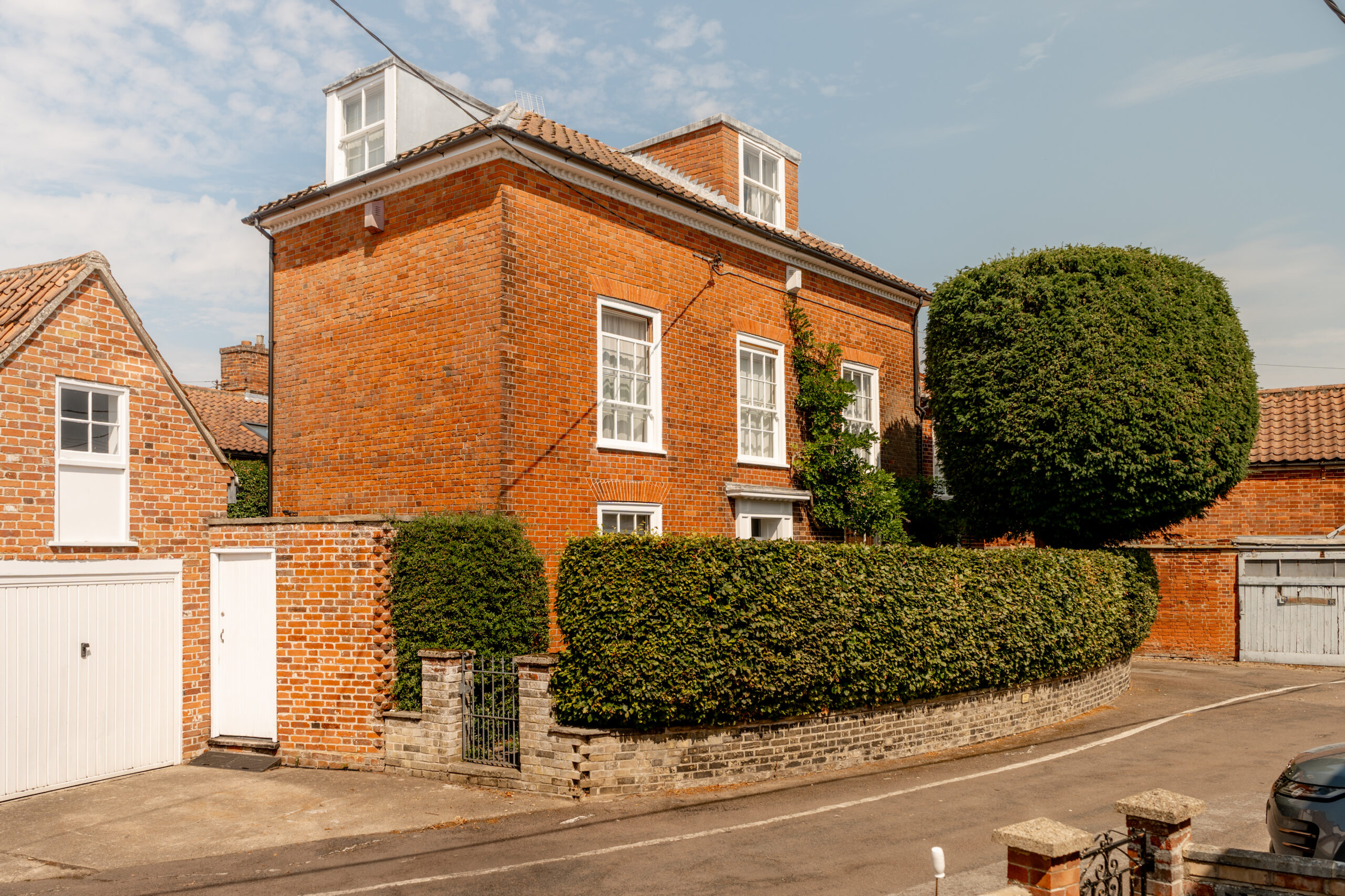 A red brick house with white-framed windows, a tall manicured hedge, and a white garage door on a quiet residential street under a partly cloudy sky.