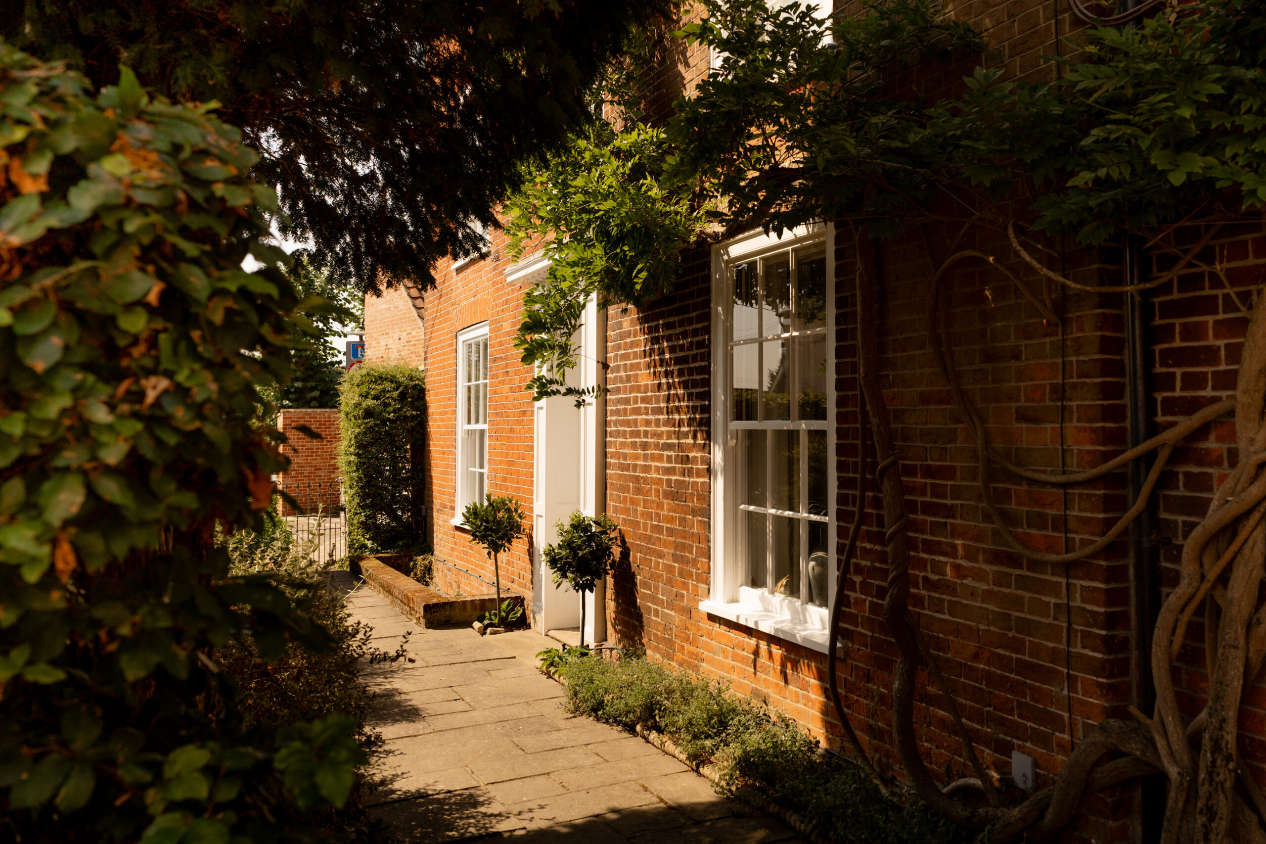 A narrow stone path runs alongside a red brick house with white-trimmed windows, lush greenery, and tree branches partially shading the walkway.