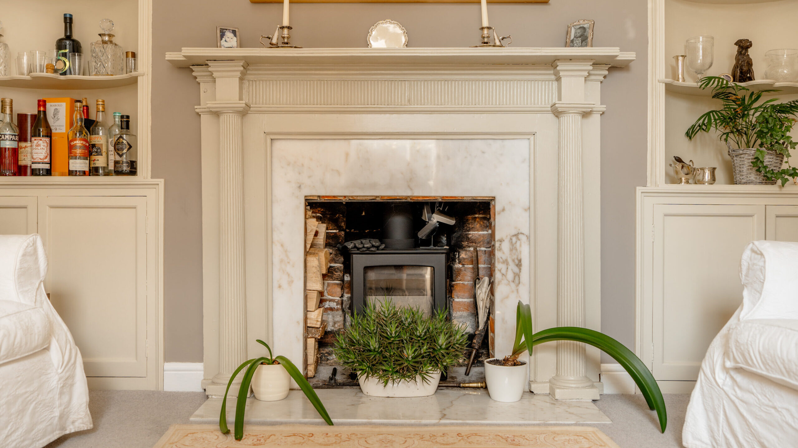 Elegant living room fireplace with marble surround, wood stove, candles, houseplants, and built-in shelves holding bottles, glassware, and decor. White armchairs and a beige rug complete the cozy, classic look.