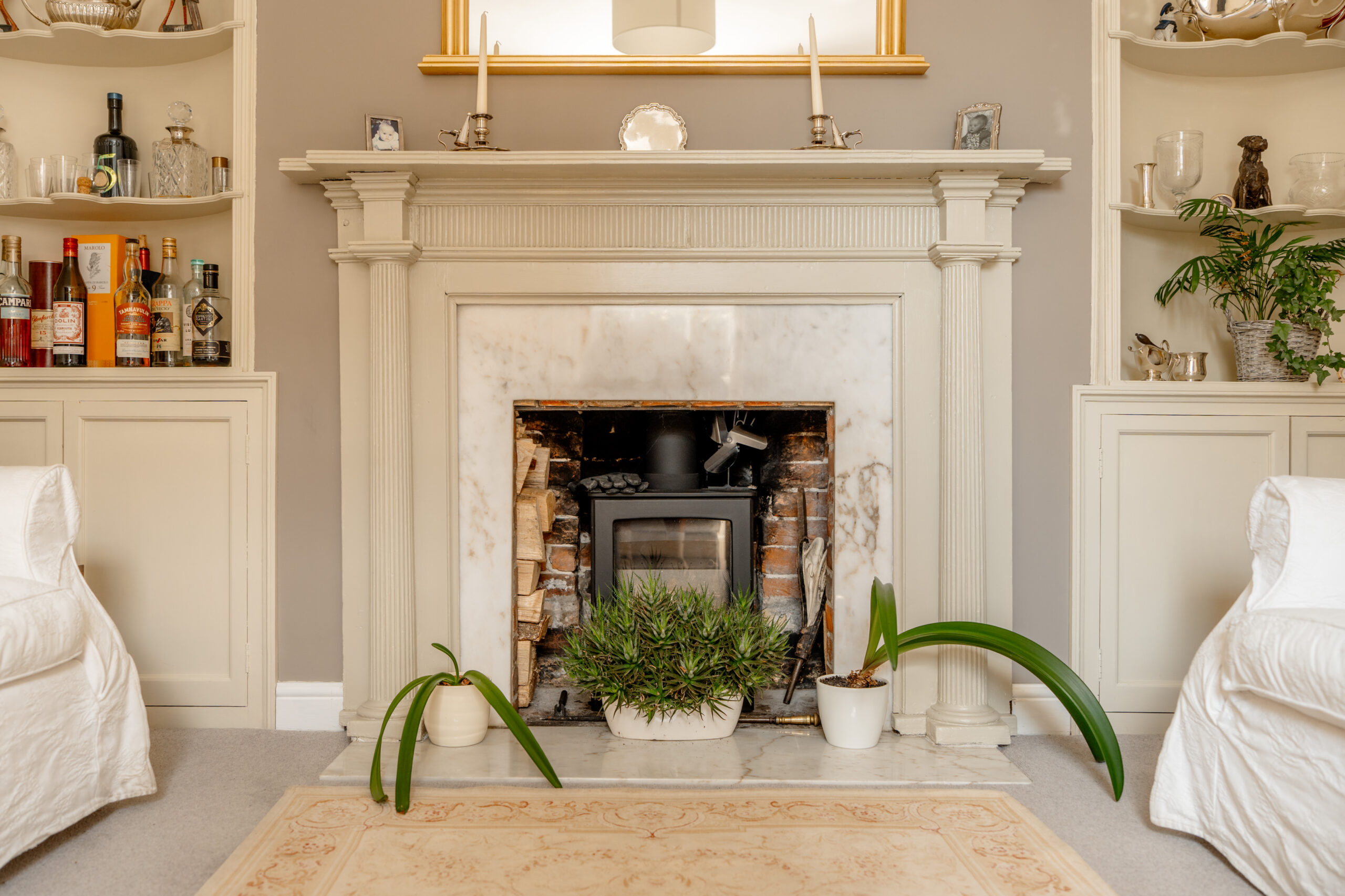 Elegant living room fireplace with marble surround, wood stove, candles, houseplants, and built-in shelves holding bottles, glassware, and decor. White armchairs and a beige rug complete the cozy, classic look.