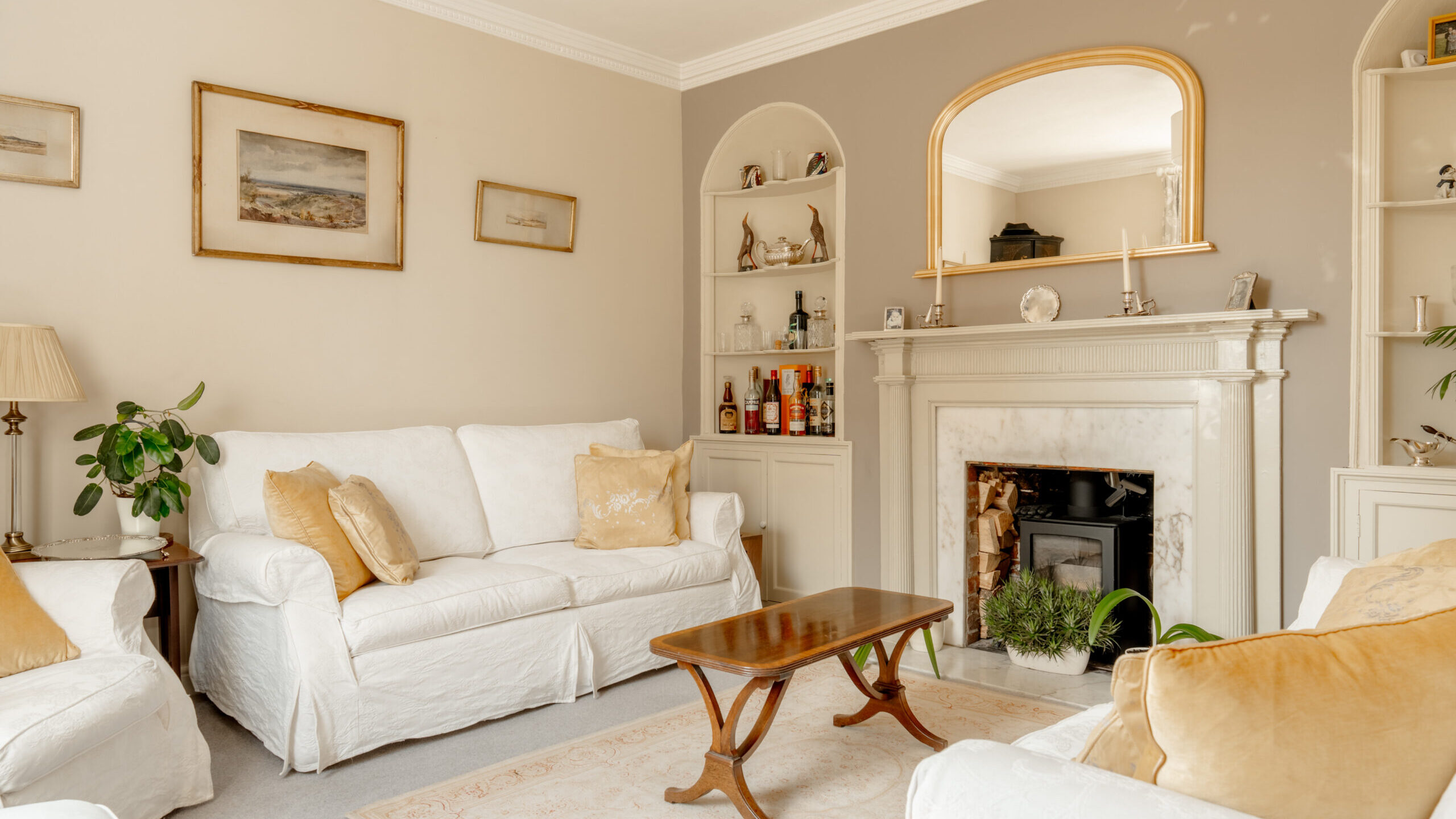 A bright living room with white sofas, a wooden coffee table, a marble fireplace, beige walls, built-in shelves, framed art, and potted plants. Neutral tones and natural light create a cozy, inviting atmosphere.