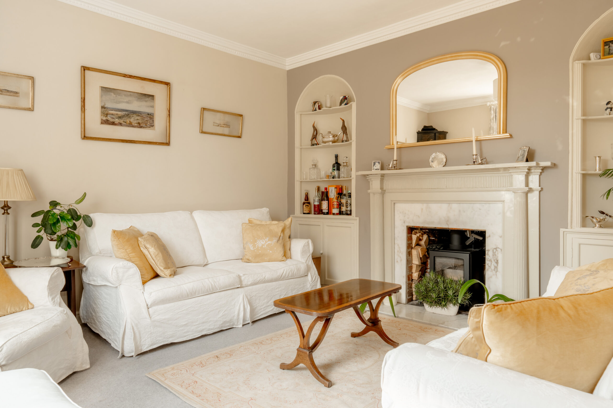 A bright living room with white sofas, a wooden coffee table, a marble fireplace, beige walls, built-in shelves, framed art, and potted plants. Neutral tones and natural light create a cozy, inviting atmosphere.