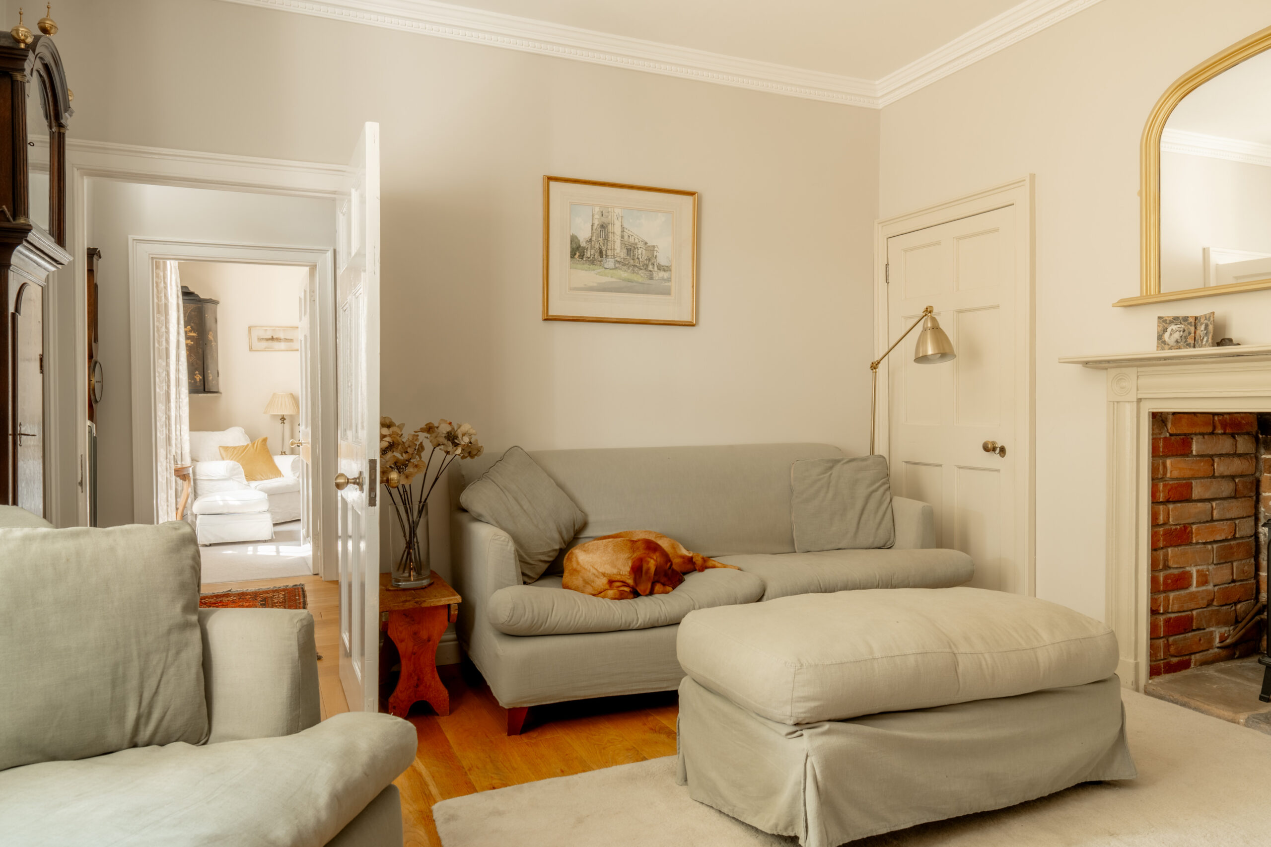 A cozy living room with light-colored sofas, a dog sleeping on one sofa, a fireplace with a mirror above it, and an open door revealing another bright room with a chair.