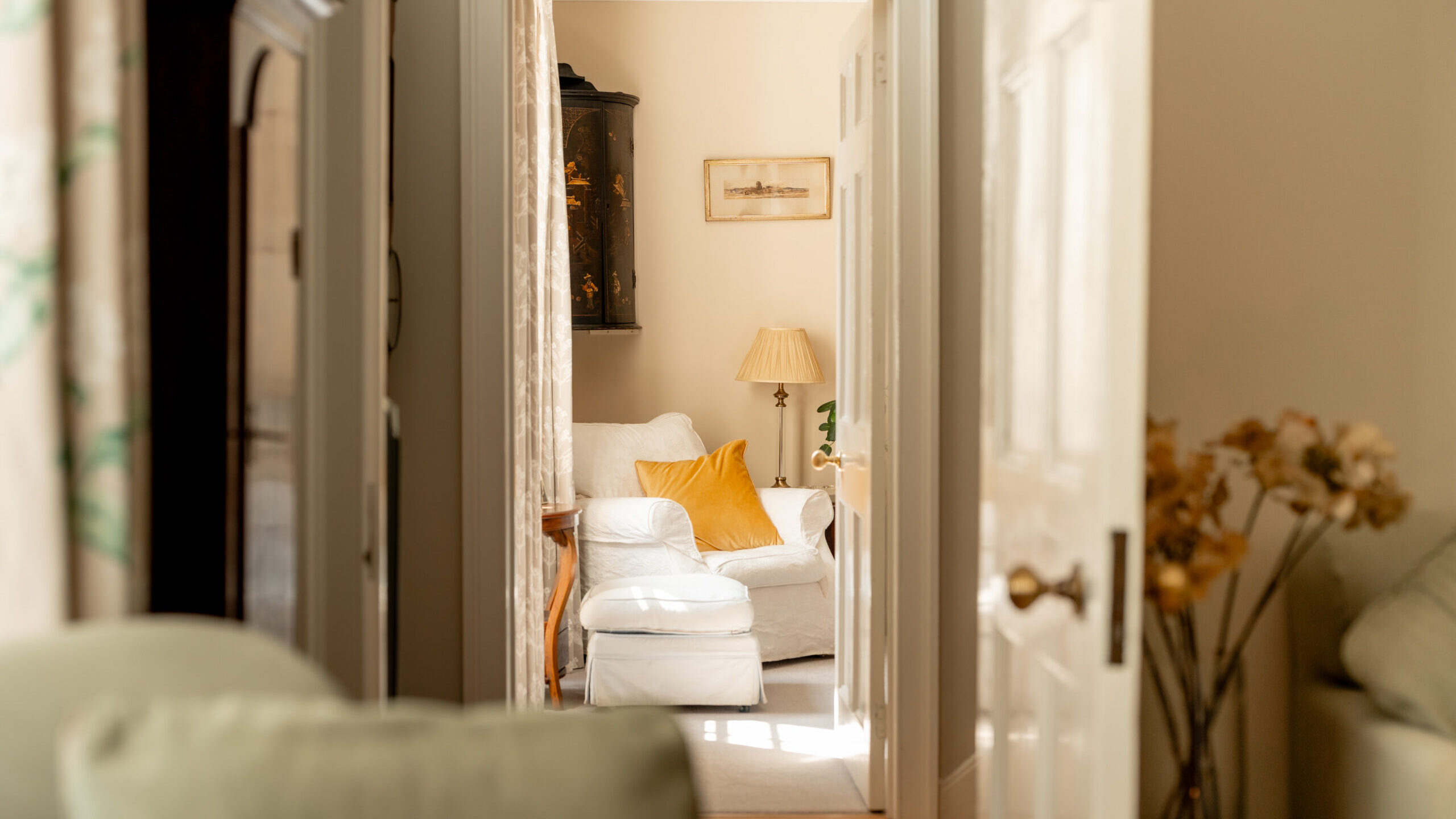 View through an open doorway into a cozy, sunlit living room with a white armchair, a yellow cushion, a lamp, framed art on the wall, and some plants and flowers.