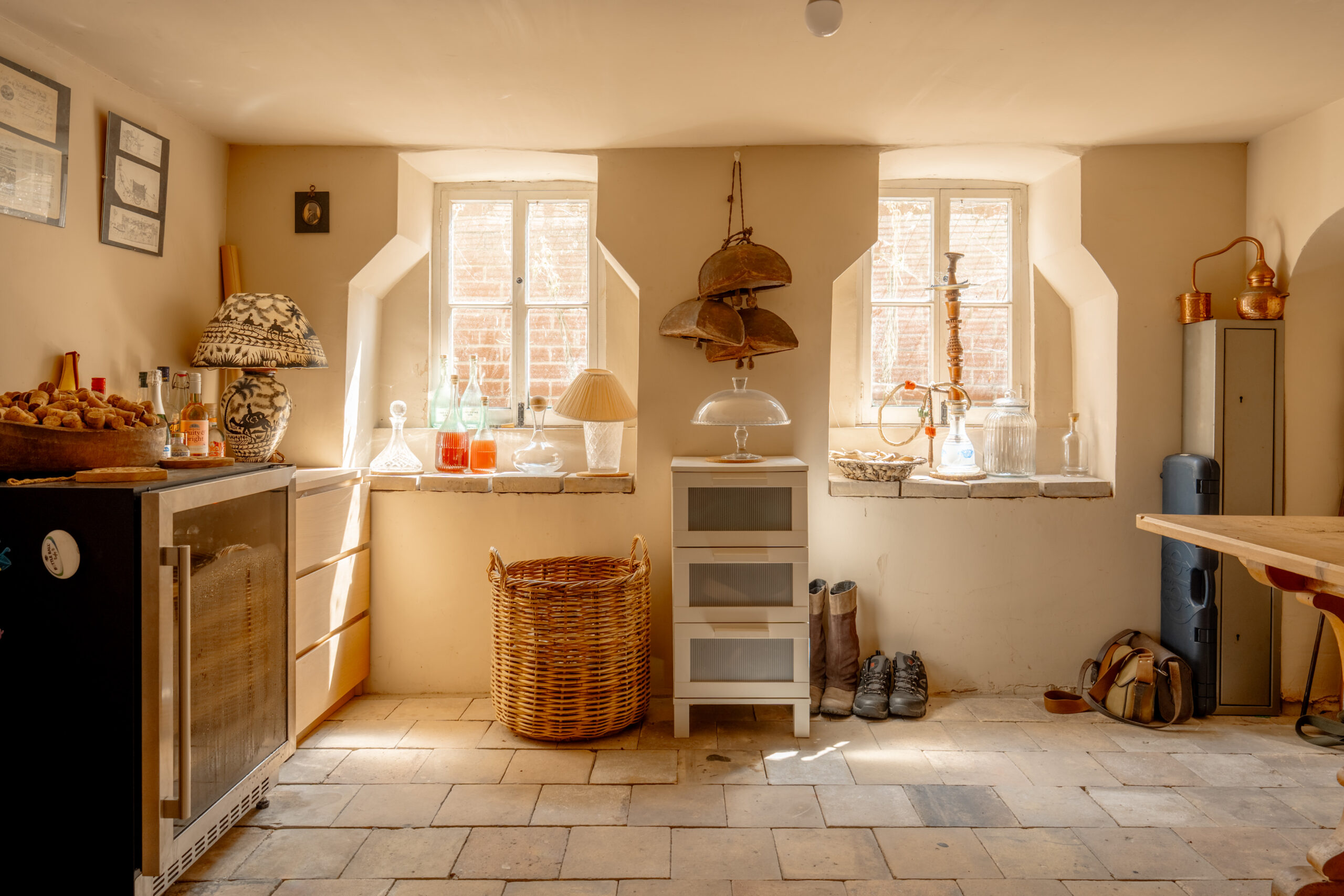 A sunlit rustic room with tiled floor, wicker basket, small cabinets, lamps, bottles, shoes, boots, and framed items on the wall, creating a cozy and organized atmosphere.