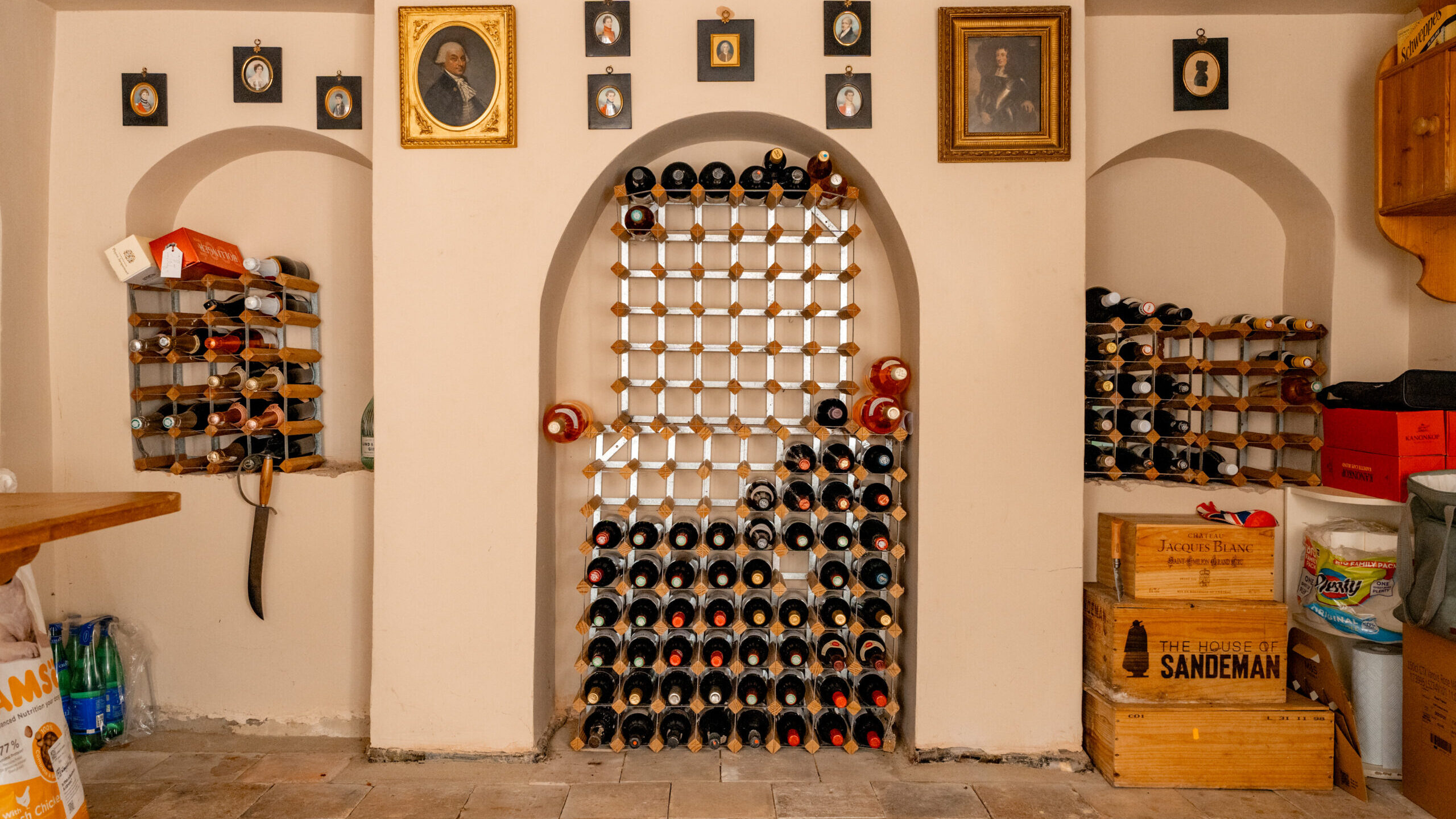 A wine cellar with a central wine rack, shelves on both sides holding bottles, framed portraits above, and wooden crates and supplies on the right. Stone floor and arched wall niches are visible.