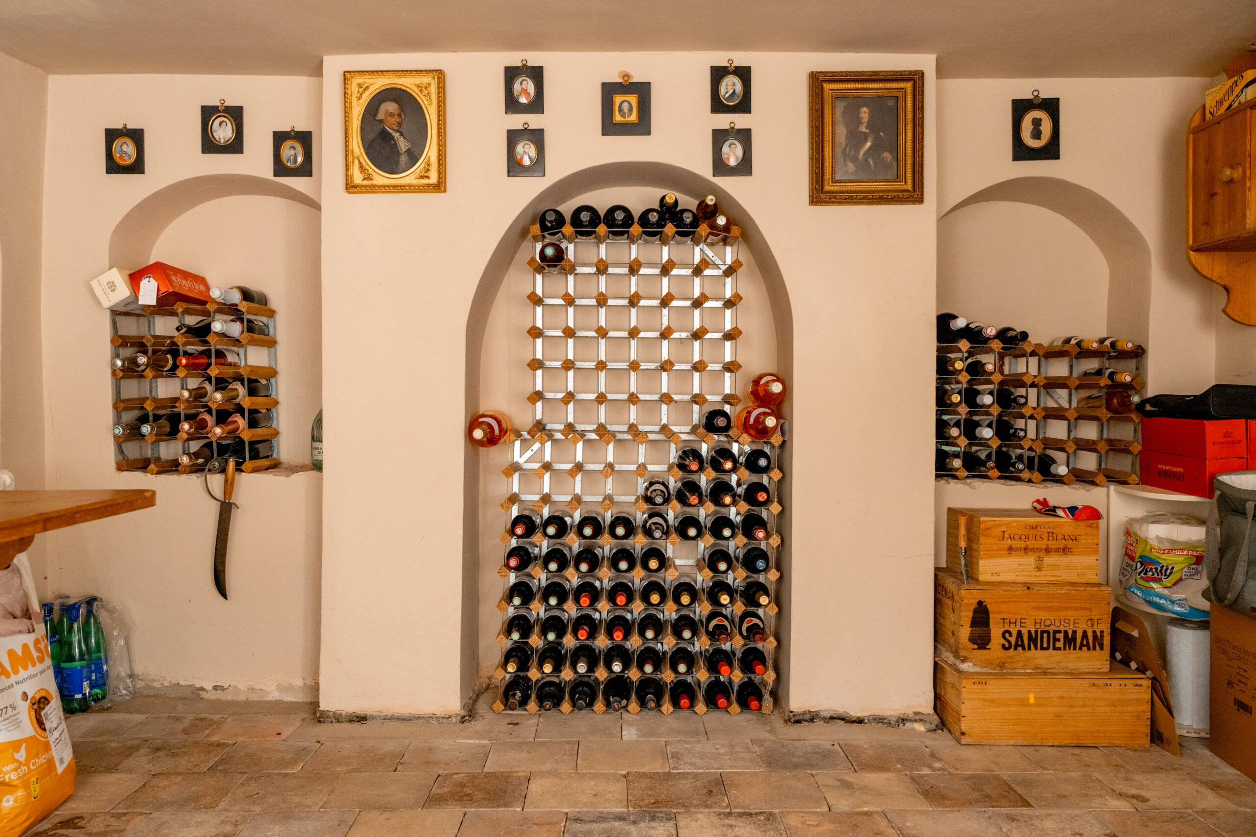 A wine cellar with a central wine rack, shelves on both sides holding bottles, framed portraits above, and wooden crates and supplies on the right. Stone floor and arched wall niches are visible.