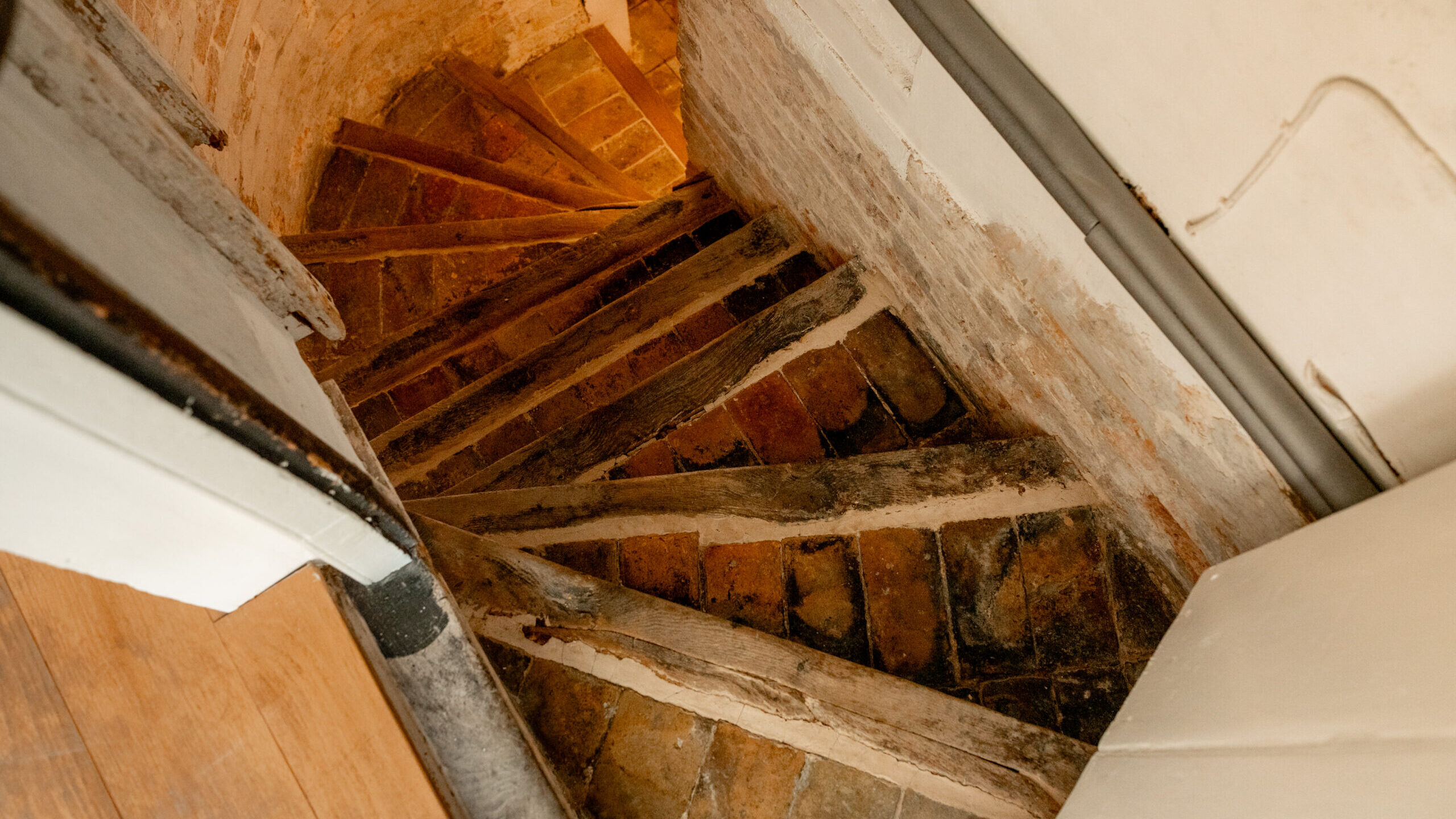 A top-down view of a rustic, curved staircase with worn wooden treads and weathered brick risers, nestled between uneven, aged white walls. The staircase descends into a dimly lit space, creating a narrow, atmospheric passage.
