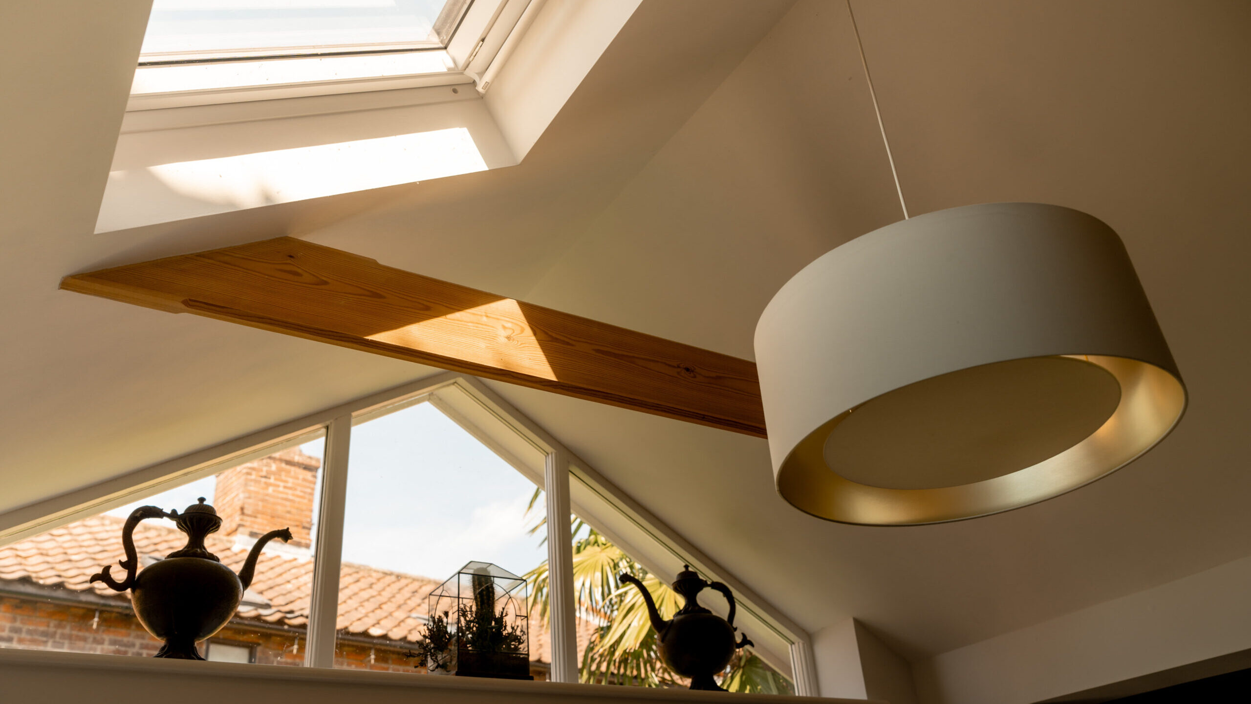 A modern pendant light hangs from a sloped ceiling with skylights. Sunlight streams in, casting shadows on the wooden beam. Two decorative vases sit on the windowsill below.