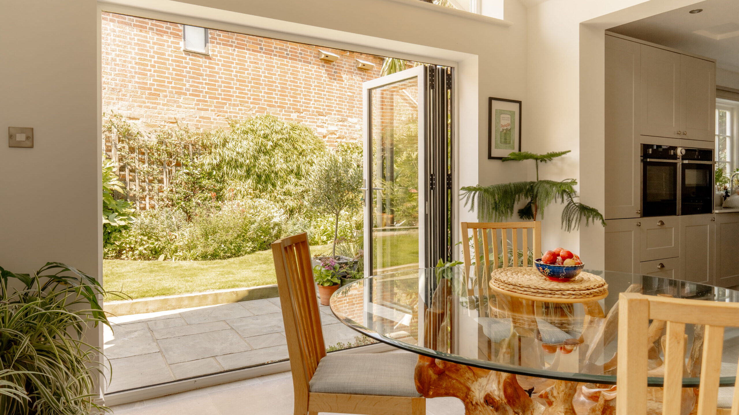 A bright kitchen dining area with a glass-topped table on a unique driftwood base. Large open bifold doors lead to a sunlit patio and lush garden featuring a brick wall, green plants, and a small lawn. A bowl of fruit sits on the table.