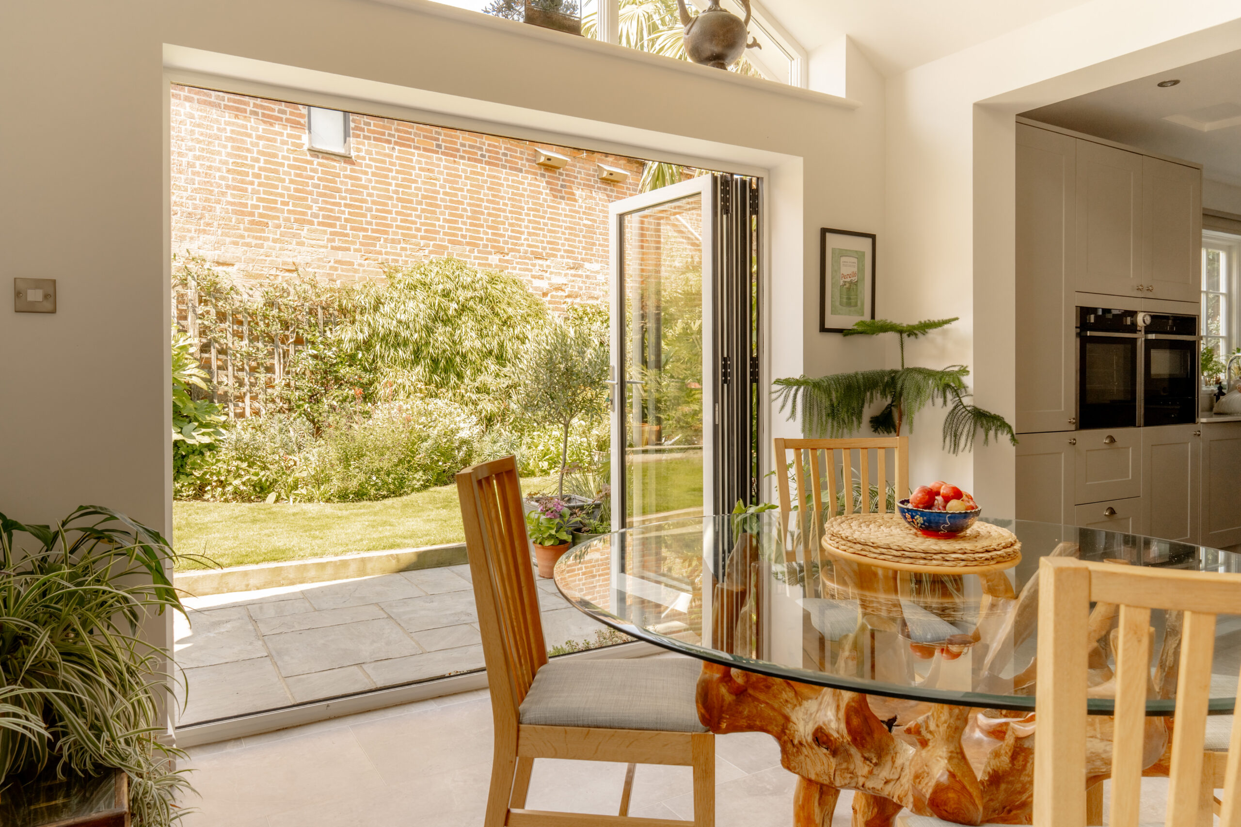 A bright kitchen dining area with a glass-topped table on a unique driftwood base. Large open bifold doors lead to a sunlit patio and lush garden featuring a brick wall, green plants, and a small lawn. A bowl of fruit sits on the table.