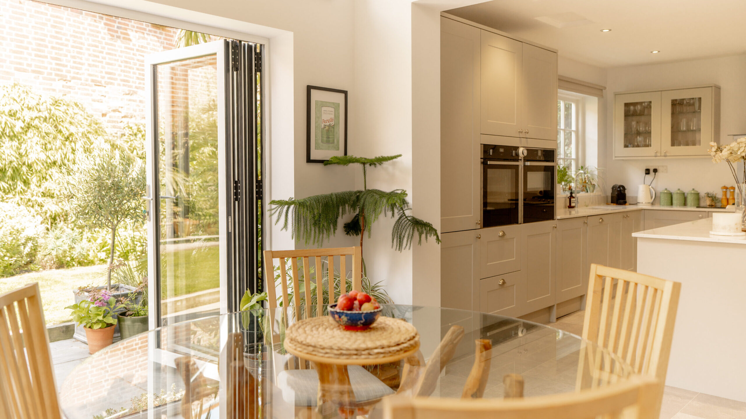 Bright kitchen and dining area with wooden chairs around a glass table, a bowl of strawberries, light cabinets, and an open door leading to a sunny garden with plants.