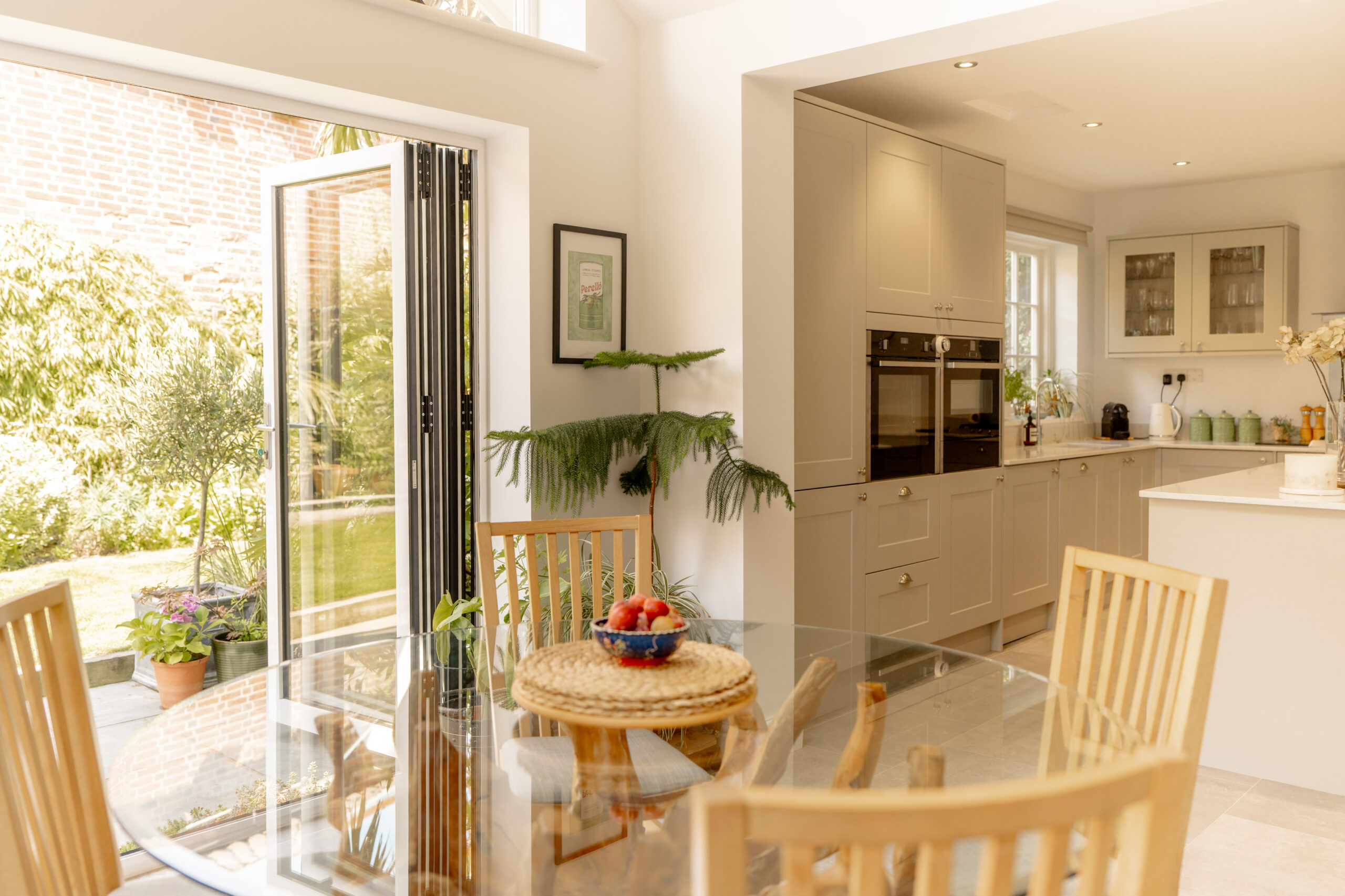 Bright kitchen and dining area with wooden chairs around a glass table, a bowl of strawberries, light cabinets, and an open door leading to a sunny garden with plants.