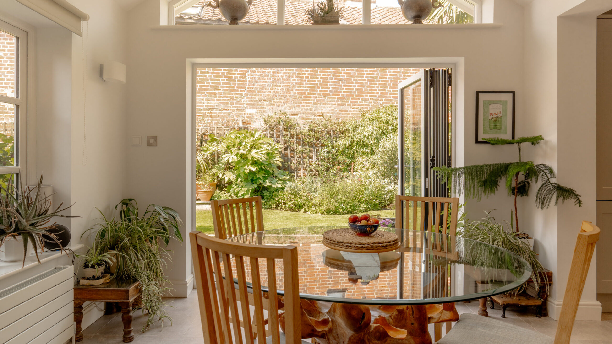 A bright, sunlit dining room features a round glass table with a unique wooden root base, surrounded by wooden chairs. Folding glass doors open to a lush garden with a brick wall. Potted plants and decorative teapots add warmth to the space.