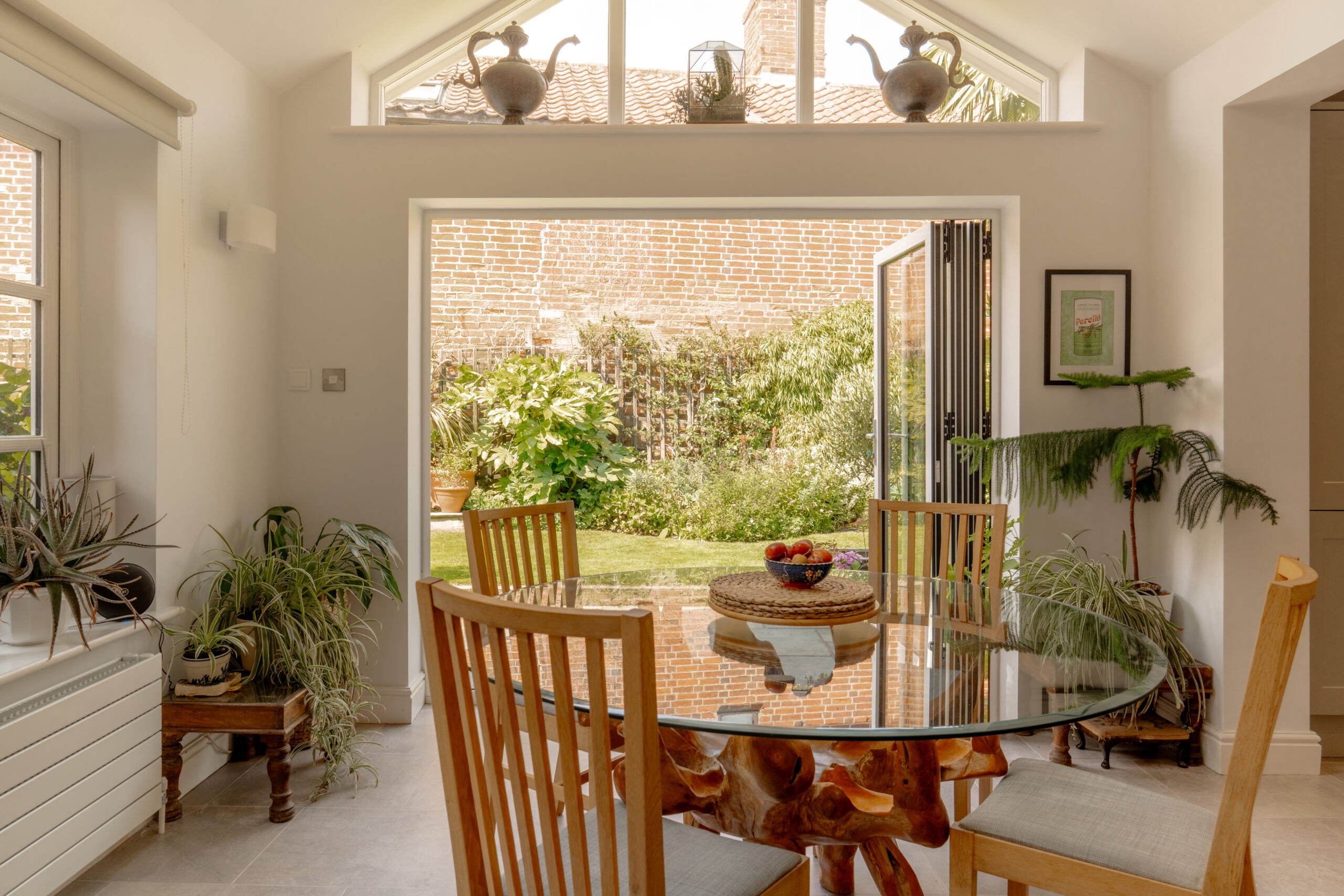 A bright, sunlit dining room features a round glass table with a unique wooden root base, surrounded by wooden chairs. Folding glass doors open to a lush garden with a brick wall. Potted plants and decorative teapots add warmth to the space.