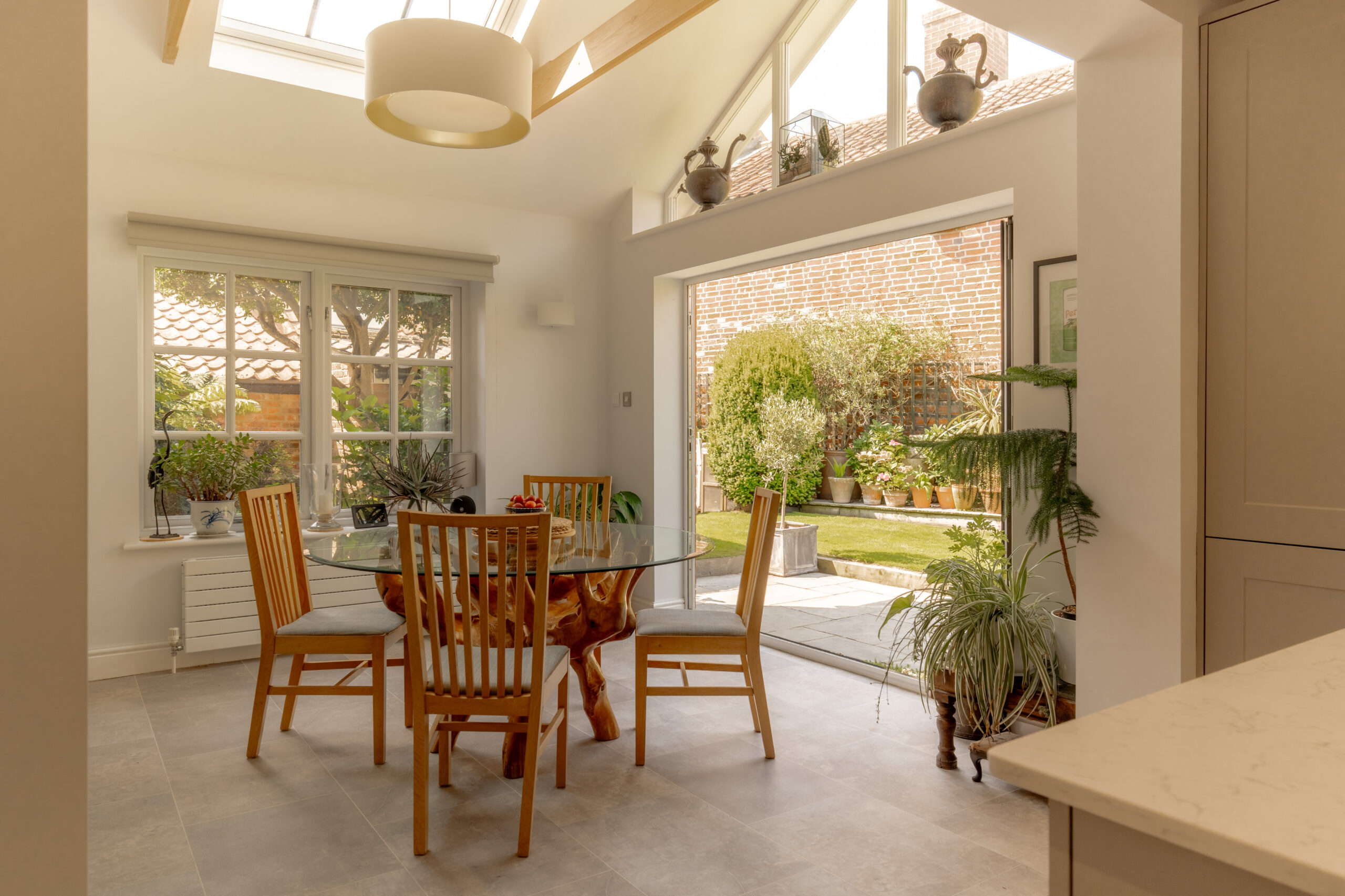 A bright dining area with a glass table and four wooden chairs. Large windows and doors open to a garden with plants. Skylights and indoor plants add to the airy, natural atmosphere.