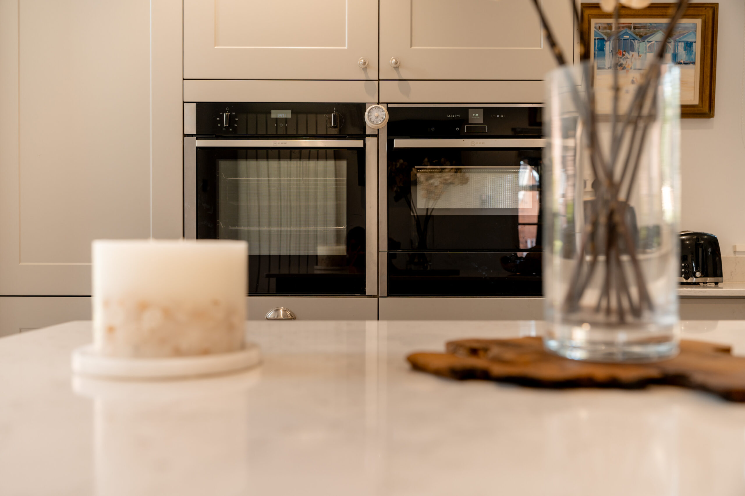 A modern kitchen with double wall ovens, a white candle on a marble counter, and a glass jar with reed diffusers on a wooden tray. A toaster and framed art are visible in the background.