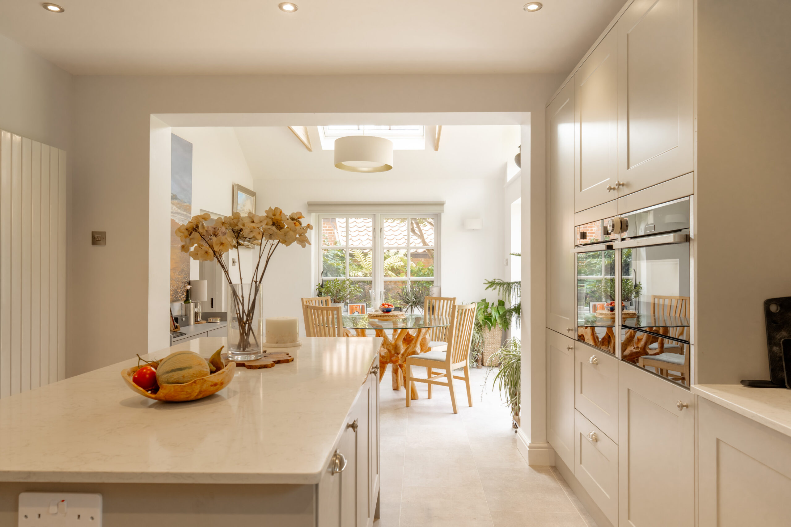Bright, modern kitchen with white cabinets and island, a fruit bowl, flowers, and sunlight streaming onto a wooden dining table with four chairs near large windows.