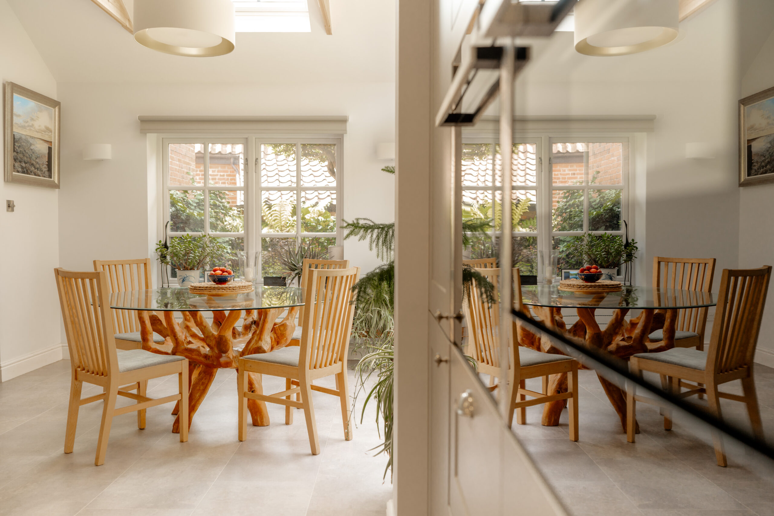 A dining room with a glass table and wooden chairs is reflected in a mirror. The table has a tree-like base, and a bowl of fruit sits on top. Large windows let in natural light and show greenery outside.