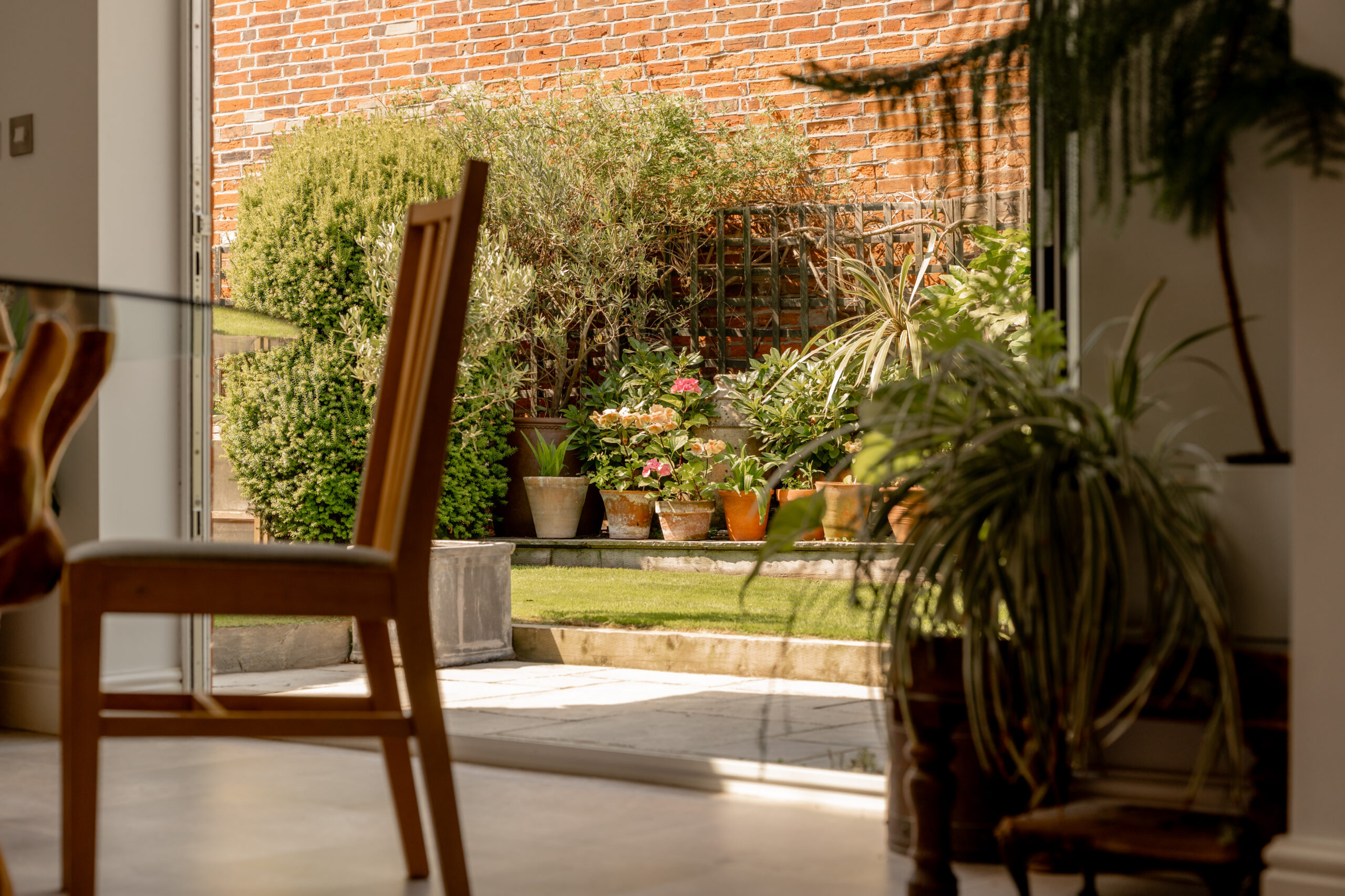A sunlit indoor dining area with wooden chairs opens to a garden patio. Outside, potted plants and green shrubs line a short wall, with a brick fence in the background.