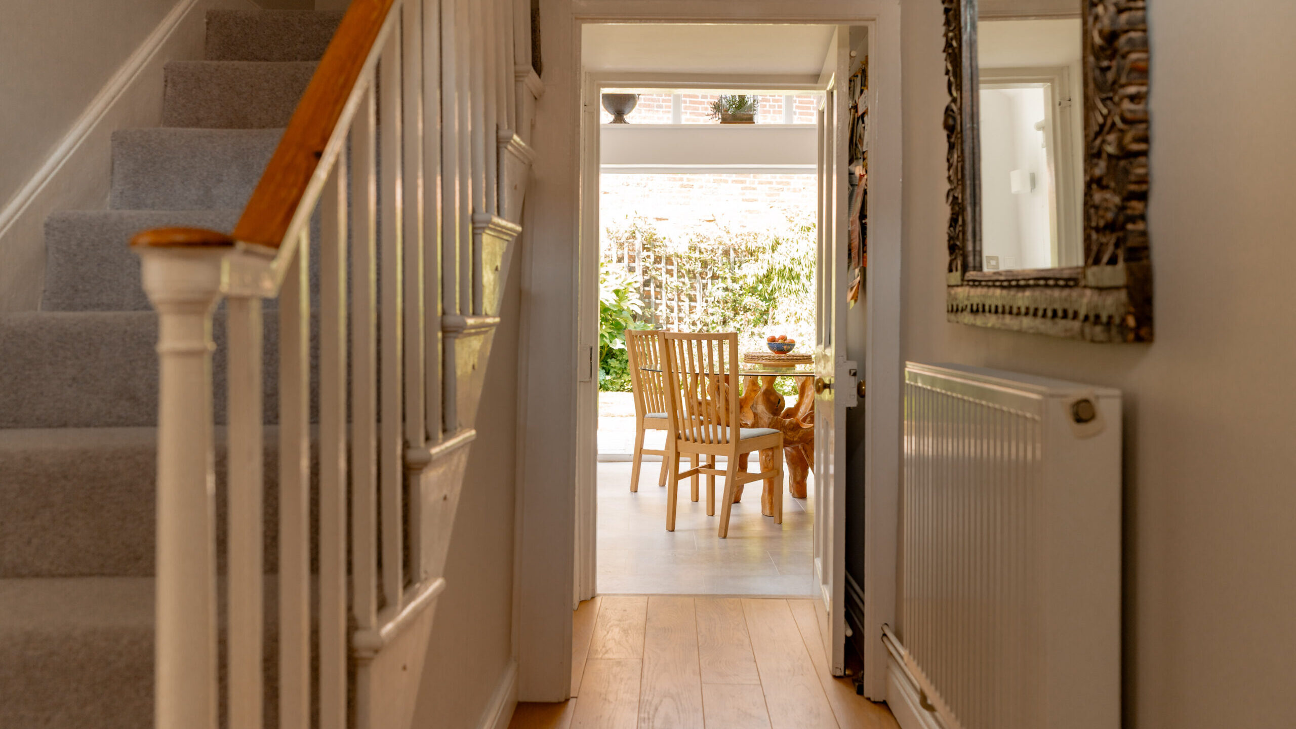 A hallway with a staircase on the left and a large mirror on the right leads to a sunlit dining area with a wooden table and chairs, and a view of greenery outside.
