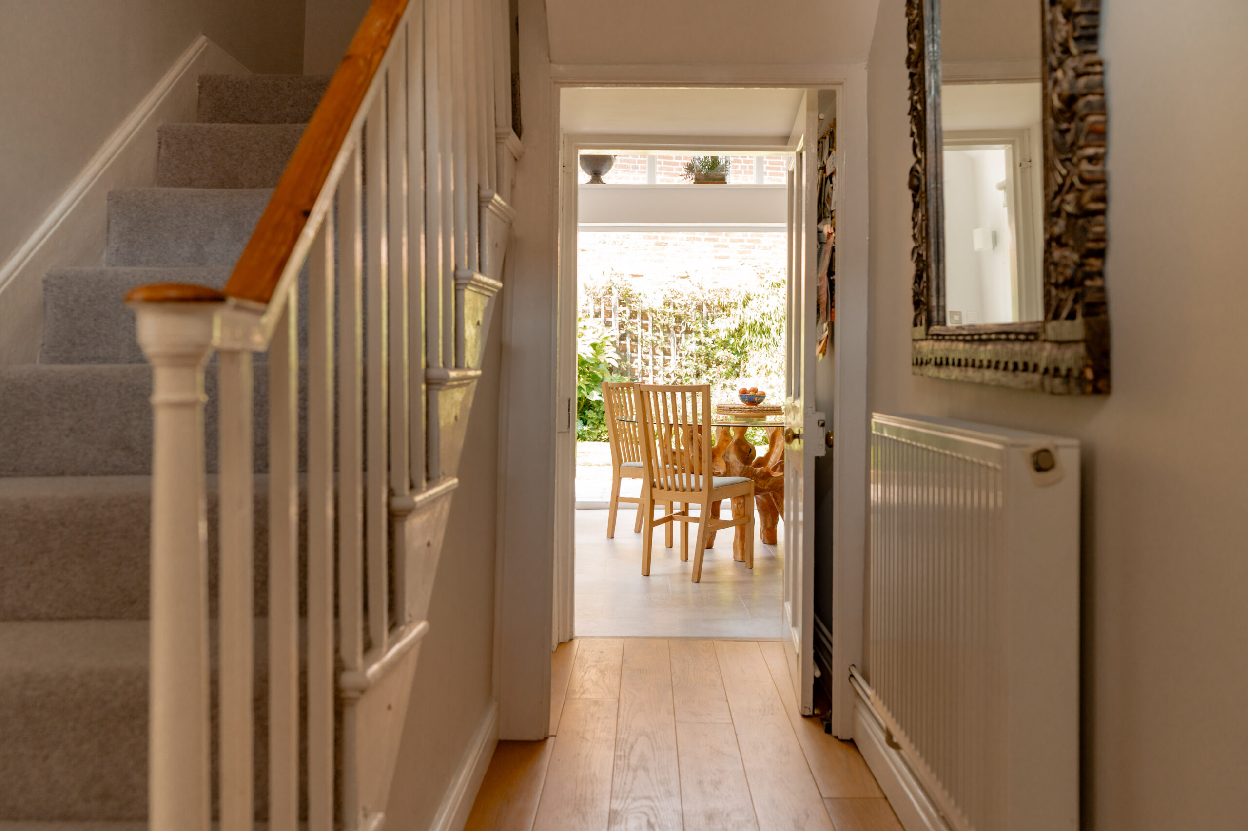 A hallway with a staircase on the left and a large mirror on the right leads to a sunlit dining area with a wooden table and chairs, and a view of greenery outside.