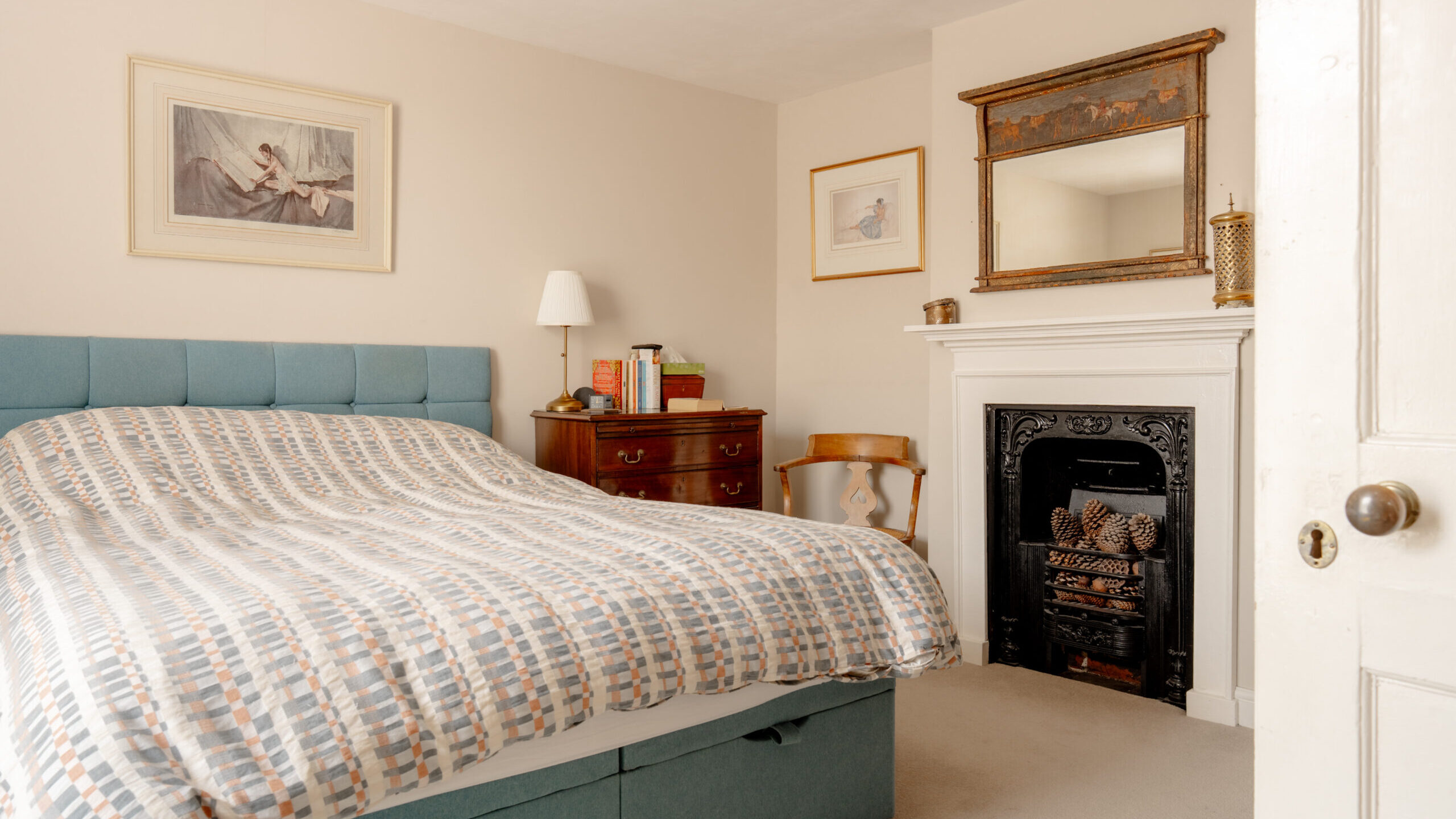 A cozy bedroom with a patterned bed, wooden furniture, a decorative fireplace, framed artwork on cream-colored walls, and a lamp on a chest of drawers.