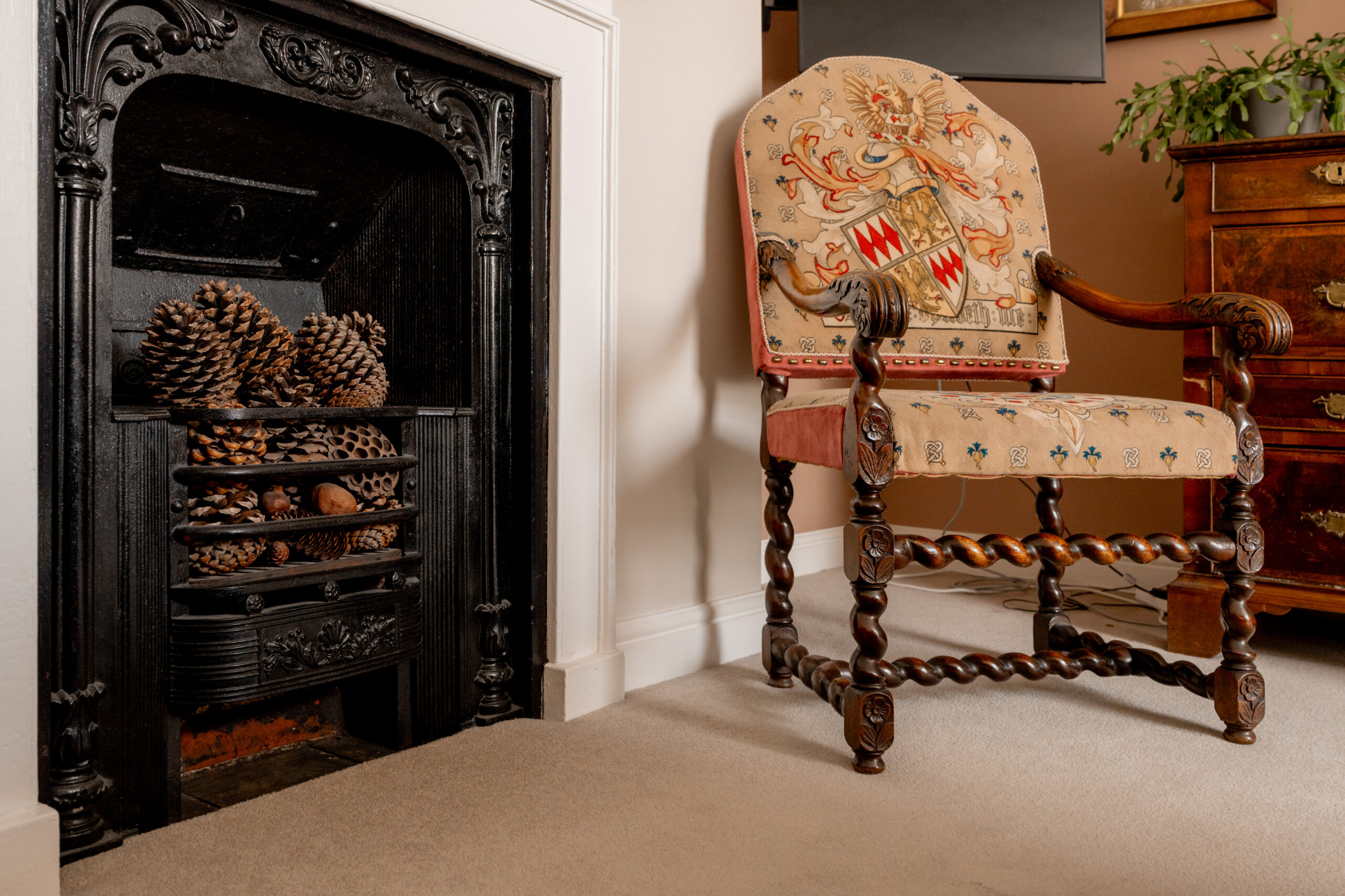 A carved wooden chair with a decorative fabric seat sits beside a black fireplace filled with pinecones in a cozy, carpeted room with a wooden dresser and a plant in the background.
