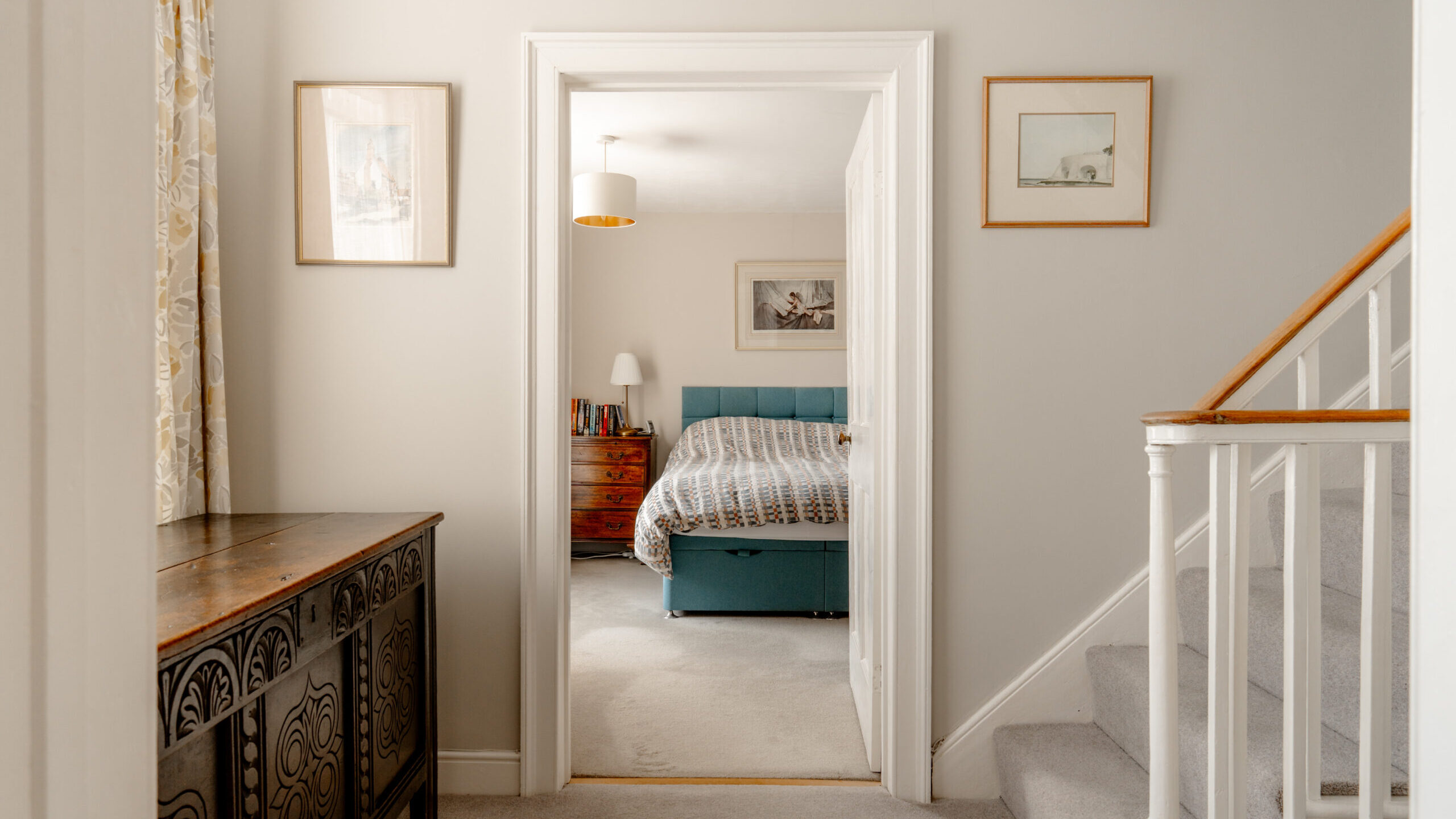View from a hallway with stairs, looking into a bedroom with a teal bed, striped bedding, a lamp, and a wooden nightstand. Light walls and framed artwork decorate the space.