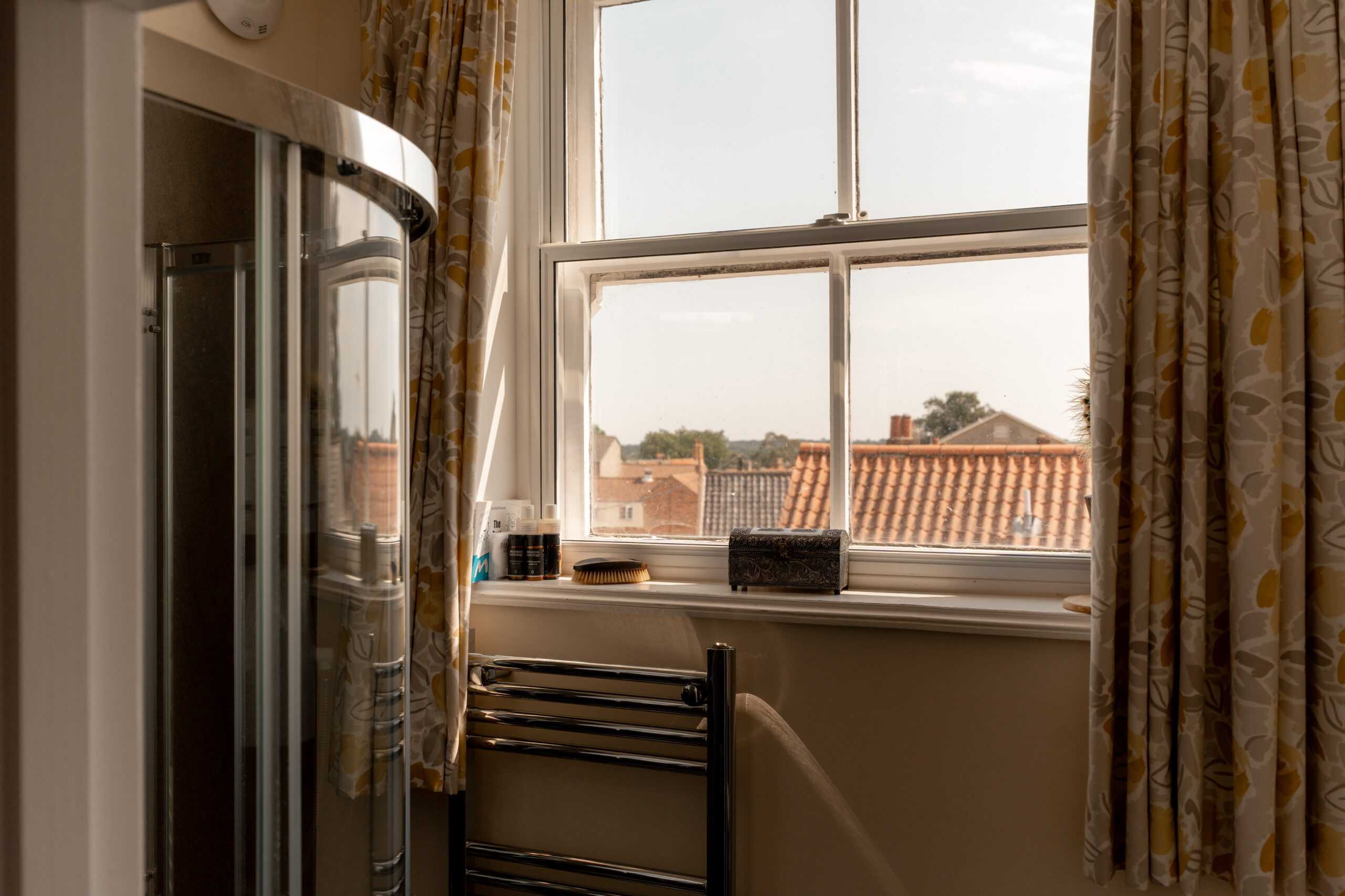 A bathroom with a glass shower, towel rack, and a window with patterned curtains. Outside, rooftops and trees are visible under a blue sky. Toiletries and a tissue box sit on the windowsill.