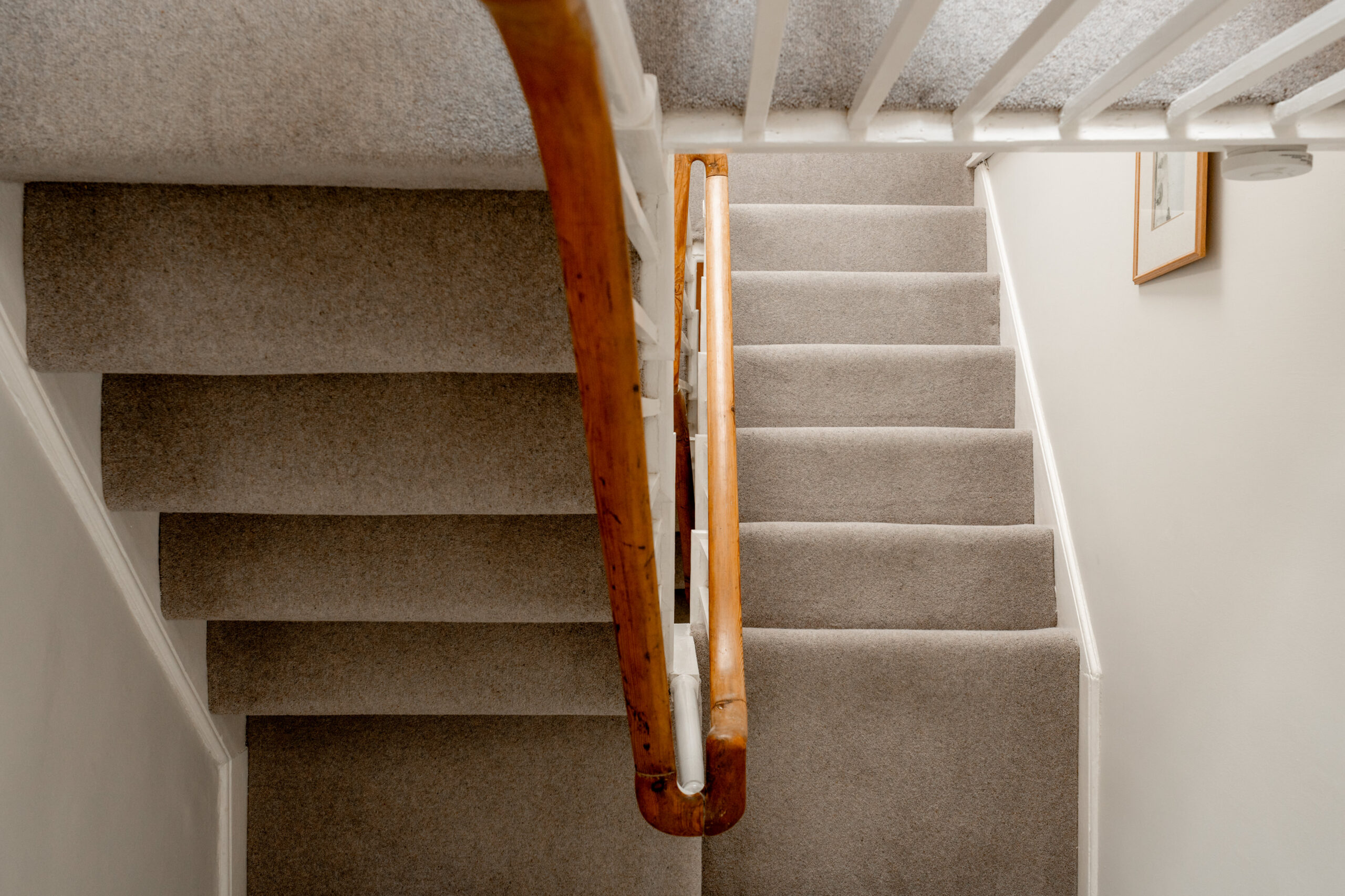 A staircase with grey carpet, wooden handrail, and white banister is viewed from above. Stairs are seen going both up and down, and a framed picture hangs on a white wall to the right.