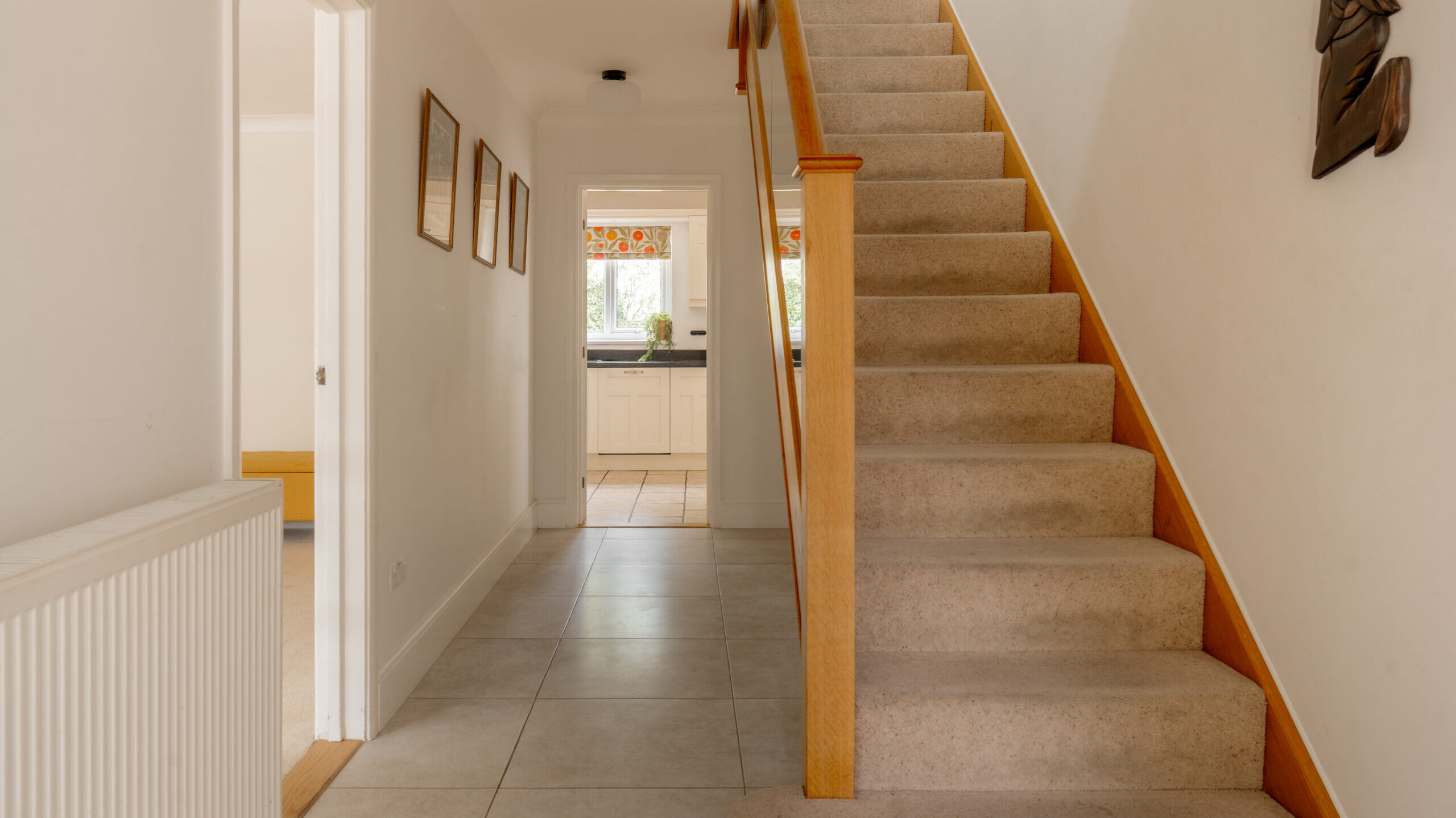 A hallway with beige carpeted stairs on the right, white tiled floor, and white walls leading to a bright kitchen at the end. Three framed pictures hang on the left wall.