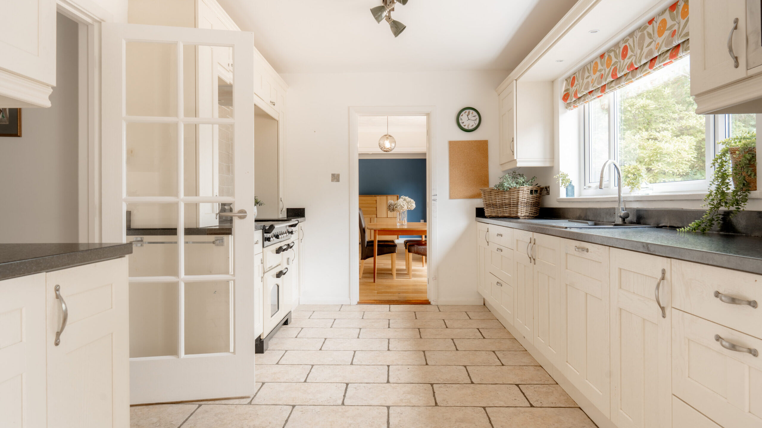 Bright kitchen with cream cabinets, black countertops, tiled floor, large window with patterned valance, and a glass door leading to a dining room with wooden furniture. Green plants are placed by the window and countertop.