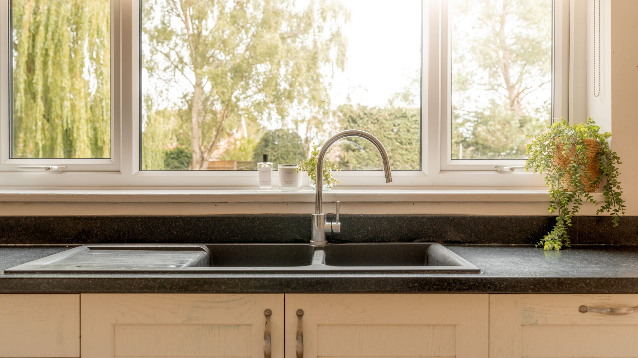 A modern kitchen sink with a silver faucet, black countertop, and potted plants on either side, positioned in front of a large window with trees visible outside.