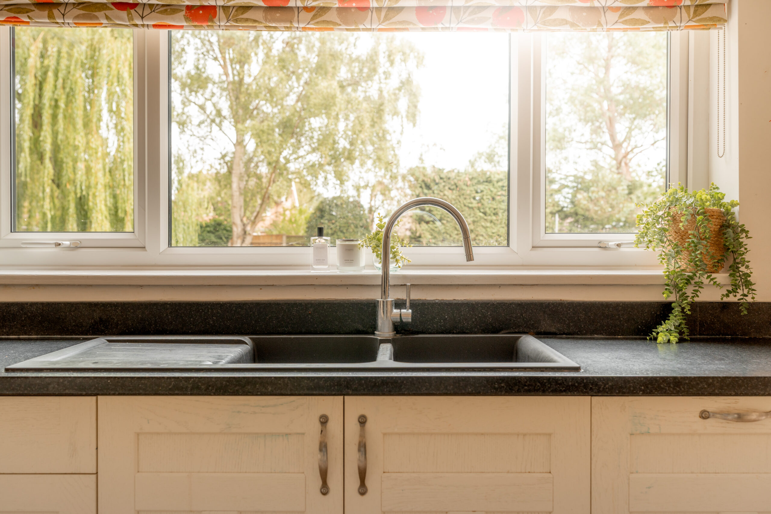 A modern kitchen sink with a silver faucet, black countertop, and potted plants on either side, positioned in front of a large window with trees visible outside.