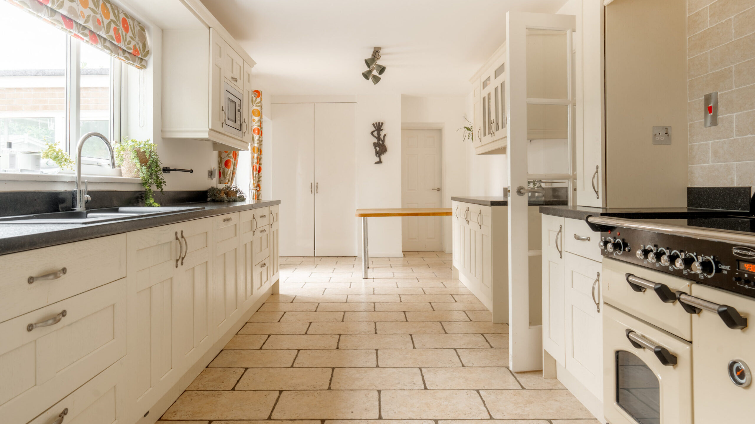 Bright, spacious kitchen with white cabinets, a large window with floral curtains, tiled floor, black countertops, a modern stove, and a small wooden table in the background.