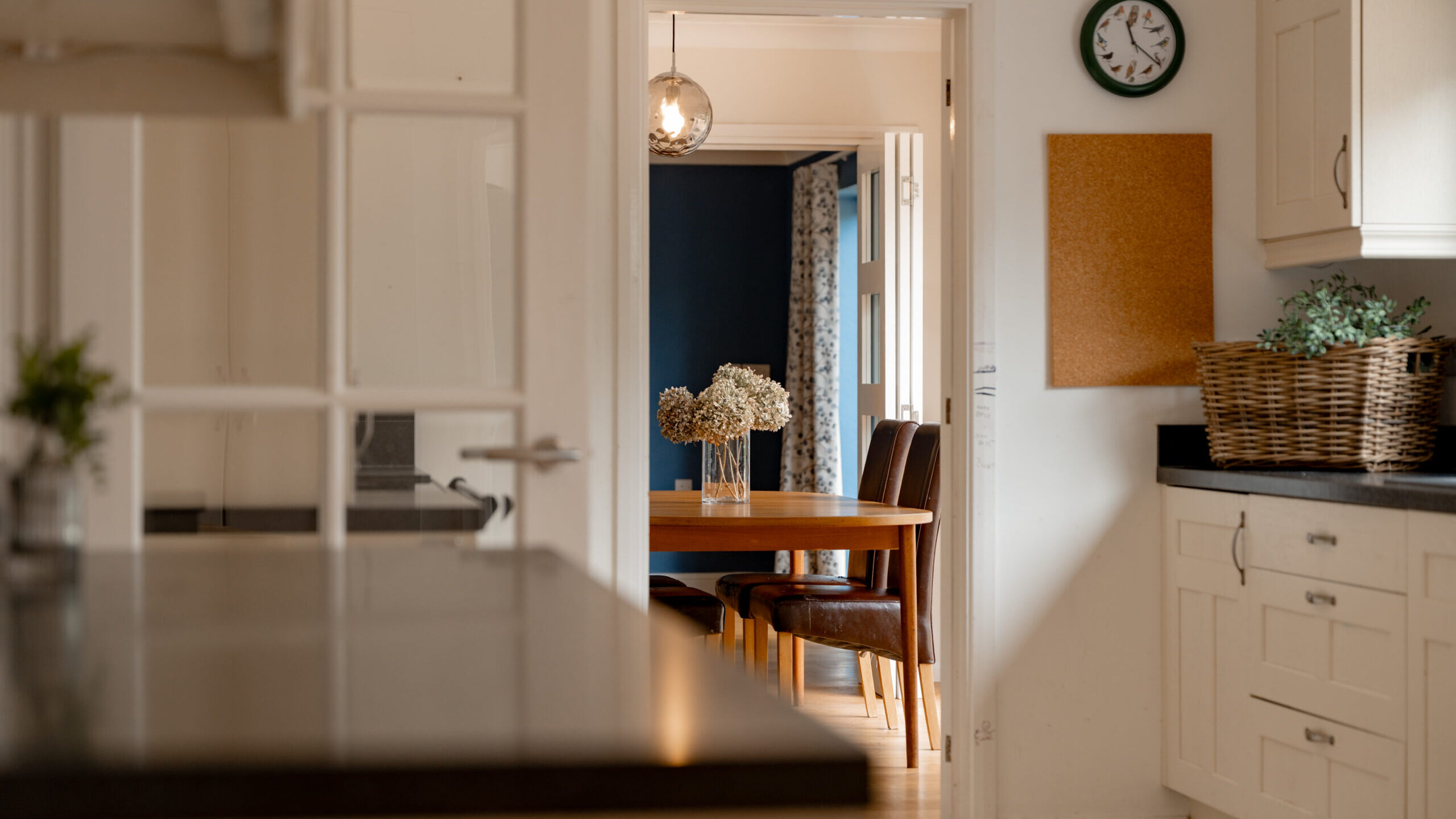 A modern kitchen with white cabinets and a black countertop opens to a dining area with a wooden table, brown chairs, and a vase of flowers. A clock, corkboard, and wicker basket are on the kitchen wall.