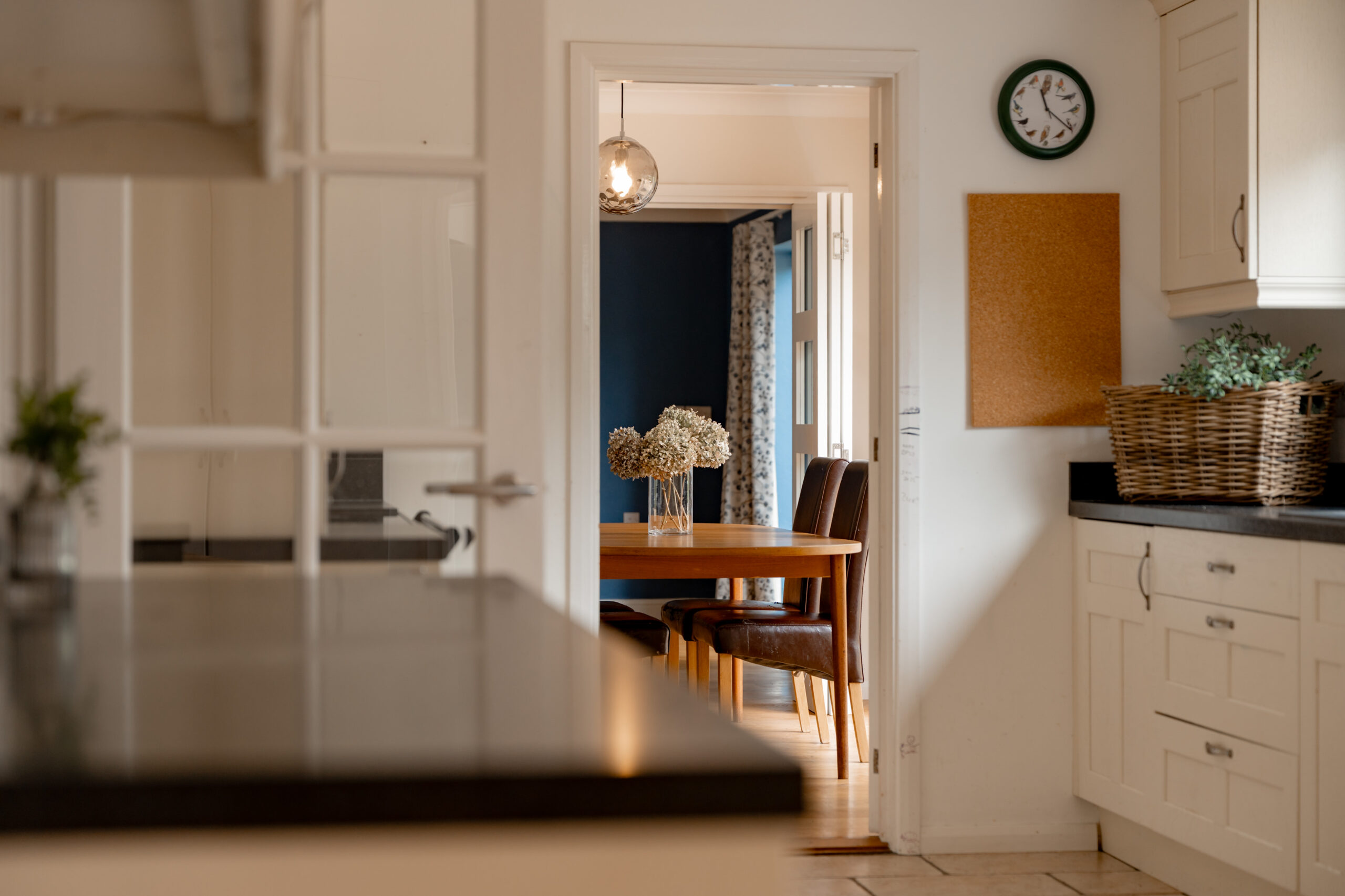 A modern kitchen with white cabinets and a black countertop opens to a dining area with a wooden table, brown chairs, and a vase of flowers. A clock, corkboard, and wicker basket are on the kitchen wall.