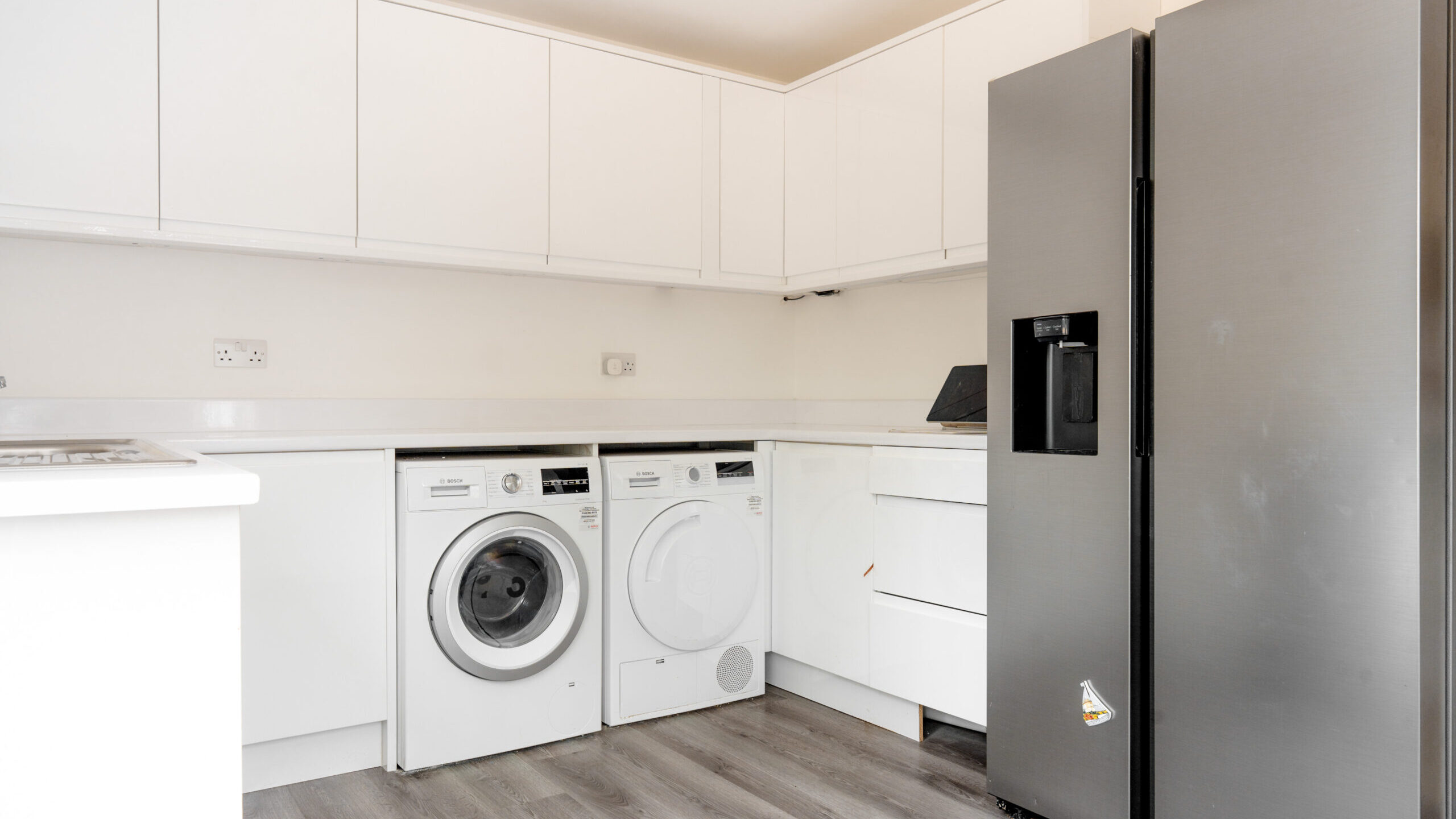 Modern kitchen with white cabinets, a washing machine, a dryer, and a large stainless steel refrigerator. The floor is wood-style laminate, and the space is clean and minimalistic.