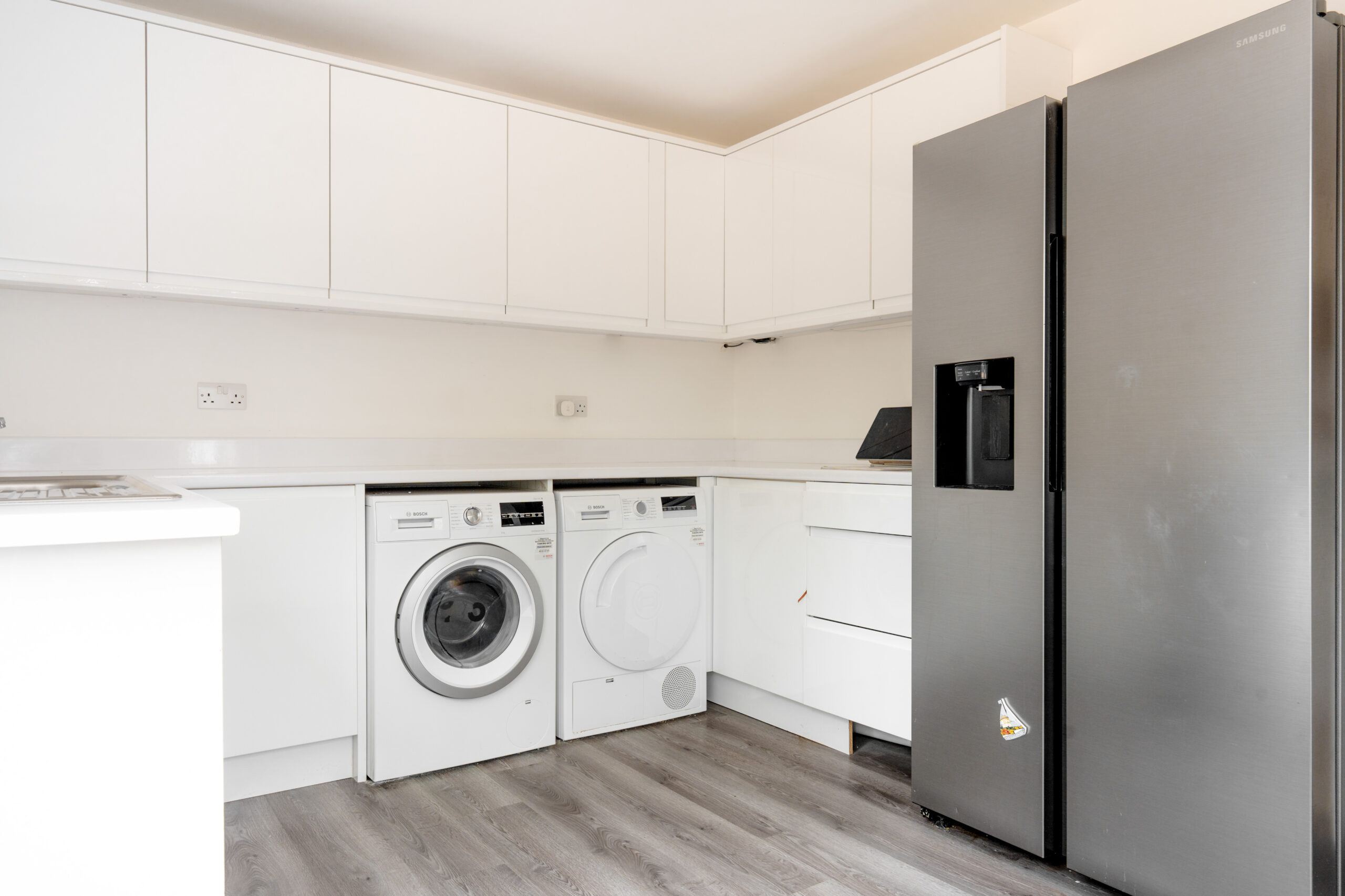 Modern kitchen with white cabinets, a washing machine, a dryer, and a large stainless steel refrigerator. The floor is wood-style laminate, and the space is clean and minimalistic.