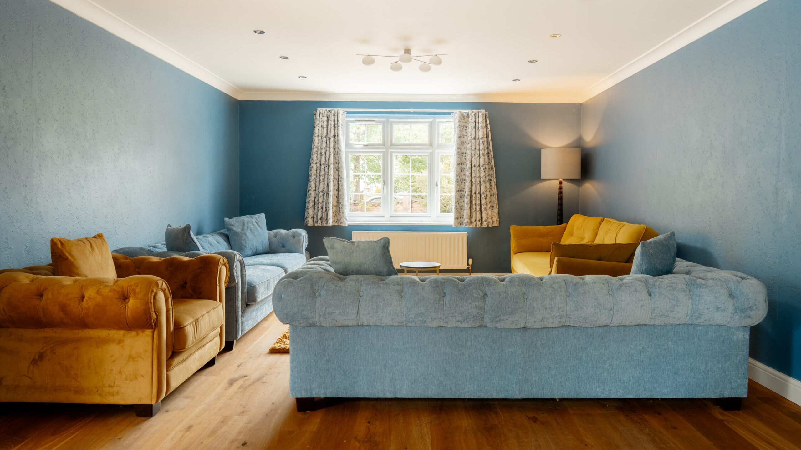 A cozy living room with blue walls, wooden floor, a large window, and three sofas—two blue and one tan. A standing lamp is in the corner, and patterned curtains are open to let natural light in.