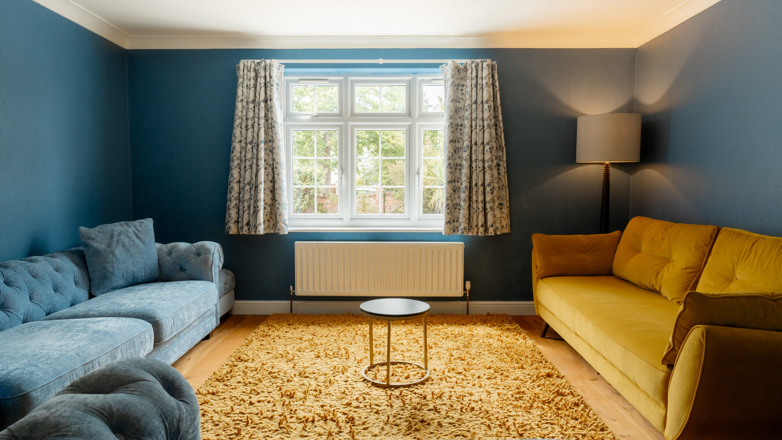 A living room with blue walls, a large window with patterned curtains, a yellow rug, a round coffee table, a blue sofa, a yellow sofa, and a floor lamp in the corner.
