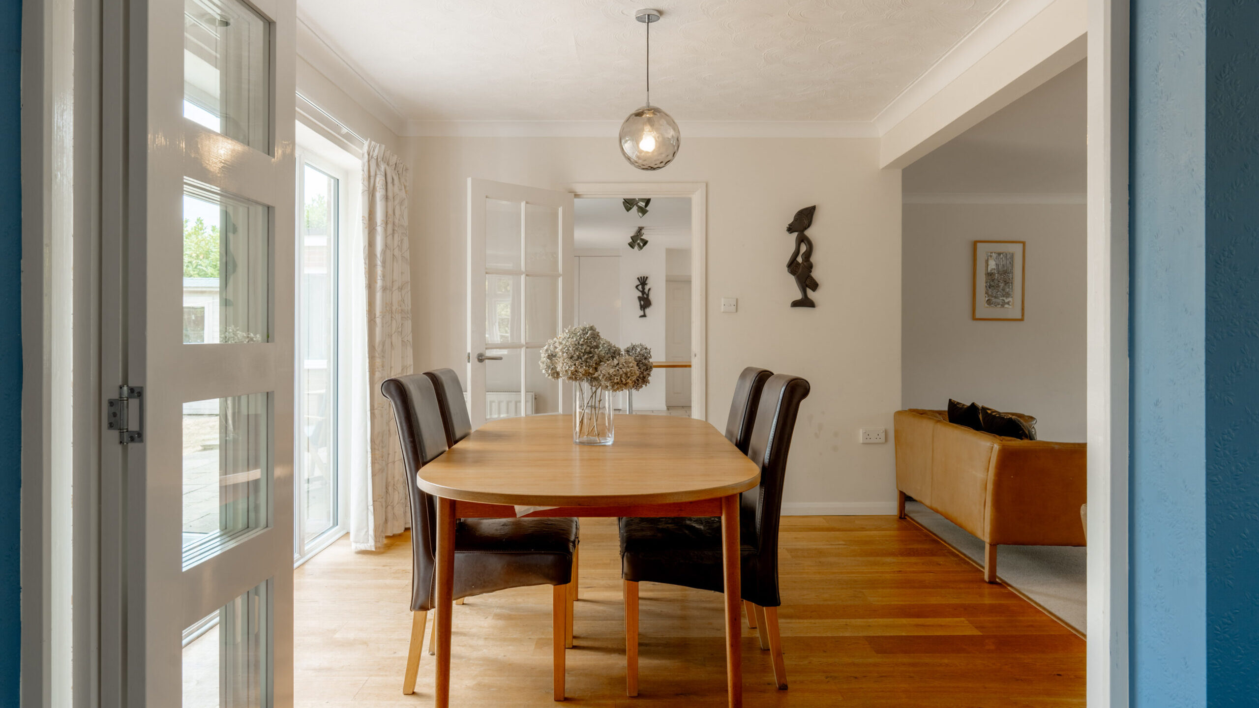 A bright dining room with a wooden table, four chairs, a vase of flowers, and glass doors leading outside. Adjacent is a living area with a brown sofa. Modern décor and hardwood floors are featured throughout.