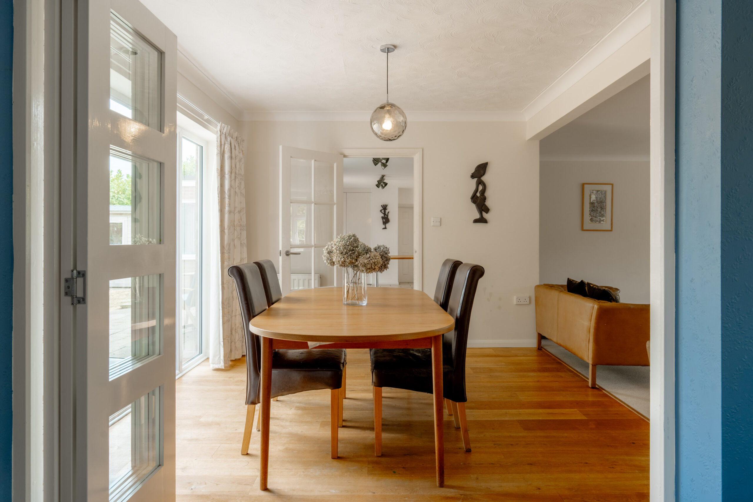 A bright dining room with a wooden table, four chairs, a vase of flowers, and glass doors leading outside. Adjacent is a living area with a brown sofa. Modern décor and hardwood floors are featured throughout.