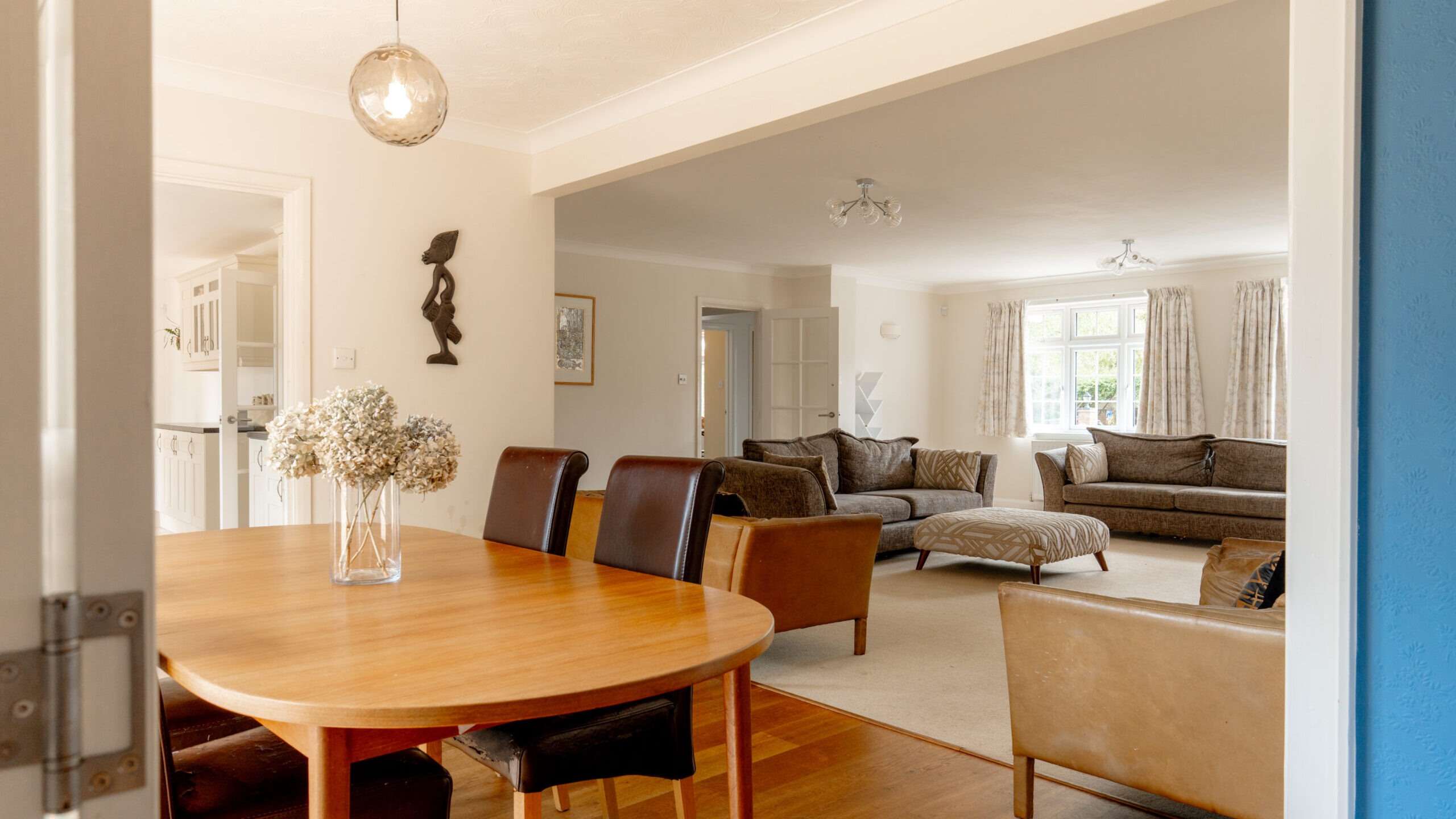 Open-plan living and dining area with a wooden table, brown chairs, beige sofas, a vase of flowers, and large windows letting in natural light.