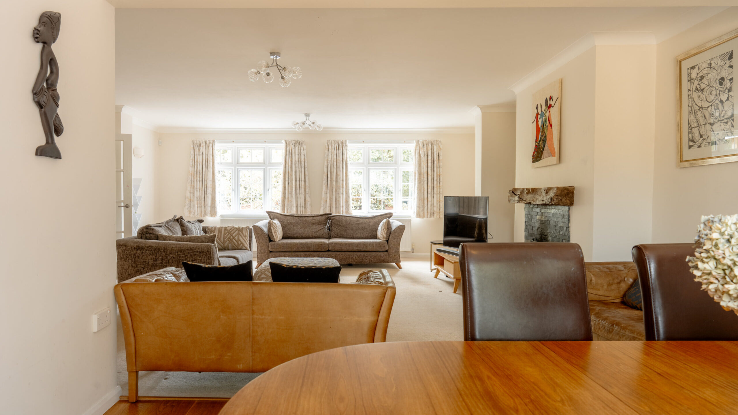 A spacious living room with large windows, beige walls, brown and tan sofas, a TV by the fireplace, and a wooden dining table in the foreground. Art and decor adorn the walls. Natural light brightens the space.