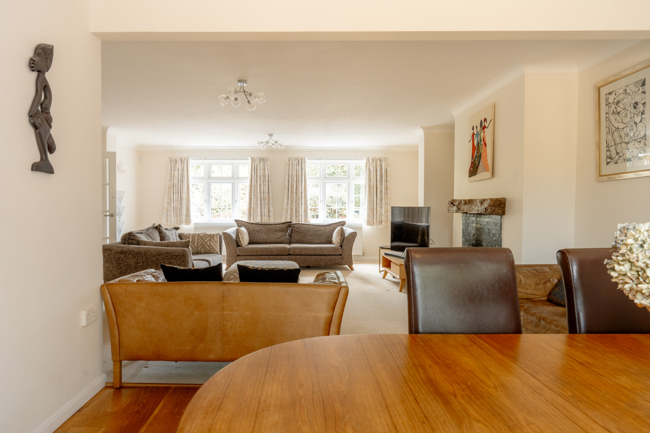 A spacious living room with large windows, beige walls, brown and tan sofas, a TV by the fireplace, and a wooden dining table in the foreground. Art and decor adorn the walls. Natural light brightens the space.